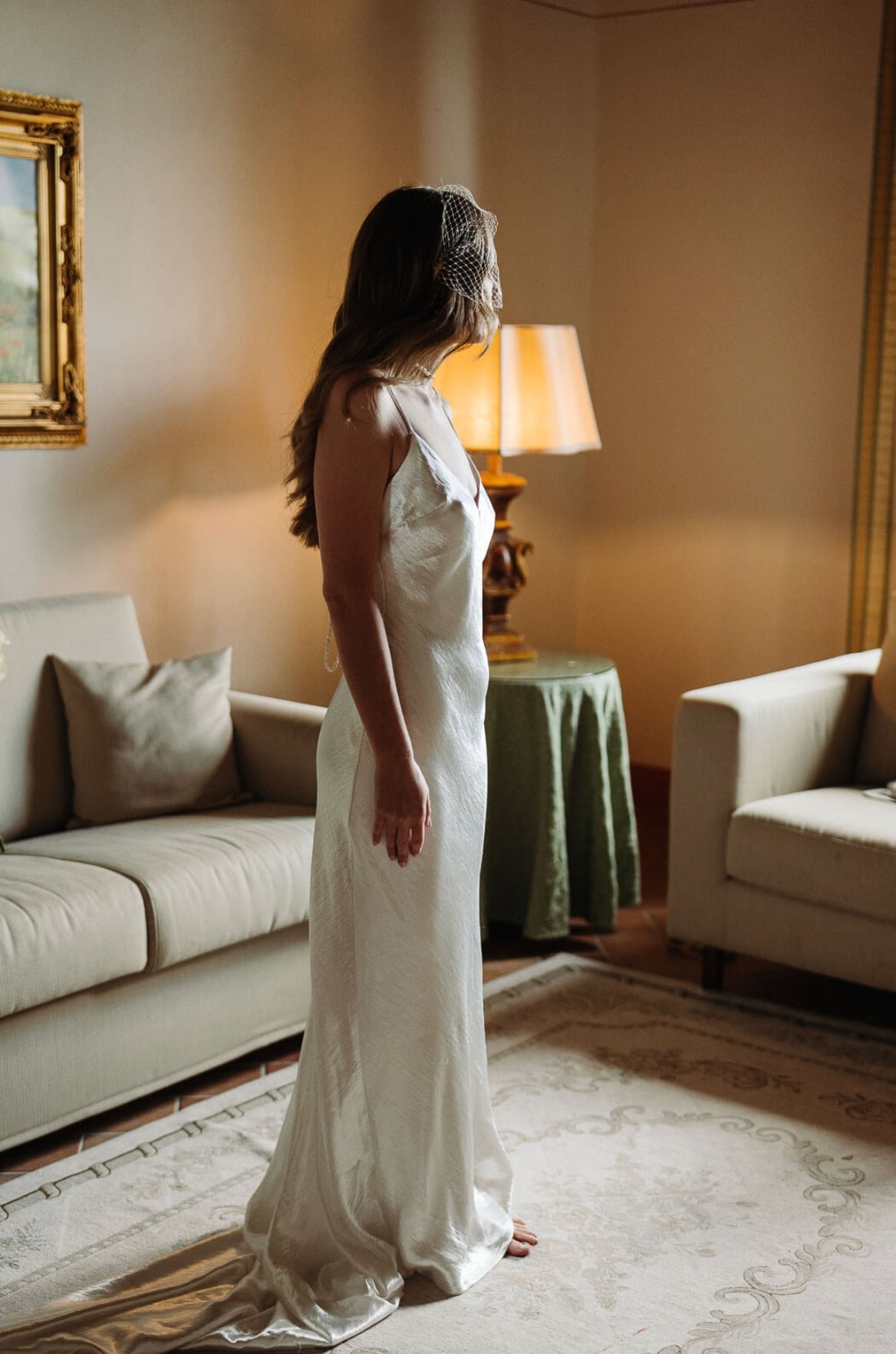 A woman in a white wedding gown with a birdcage veil standing barefoot in a living room, looking to the side. There is a beige sofa with pillows and a side table with a green tablecloth and lamp behind her.