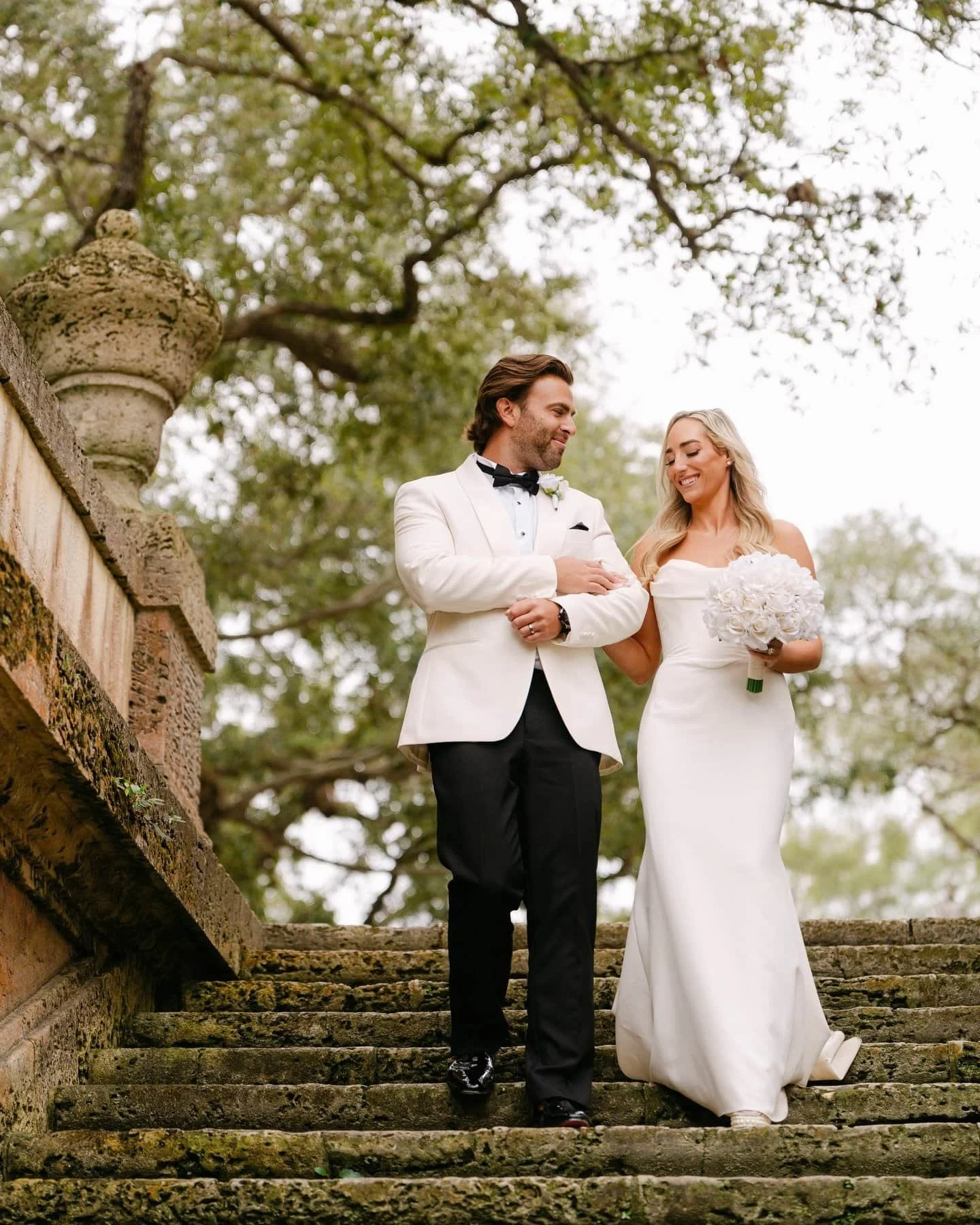 A bride and groom walking down a moss-covered stone staircase outdoors, with trees in the background. The groom is in a white tuxedo and the bride in a white wedding dress holding a bouquet of white flowers, both smiling.