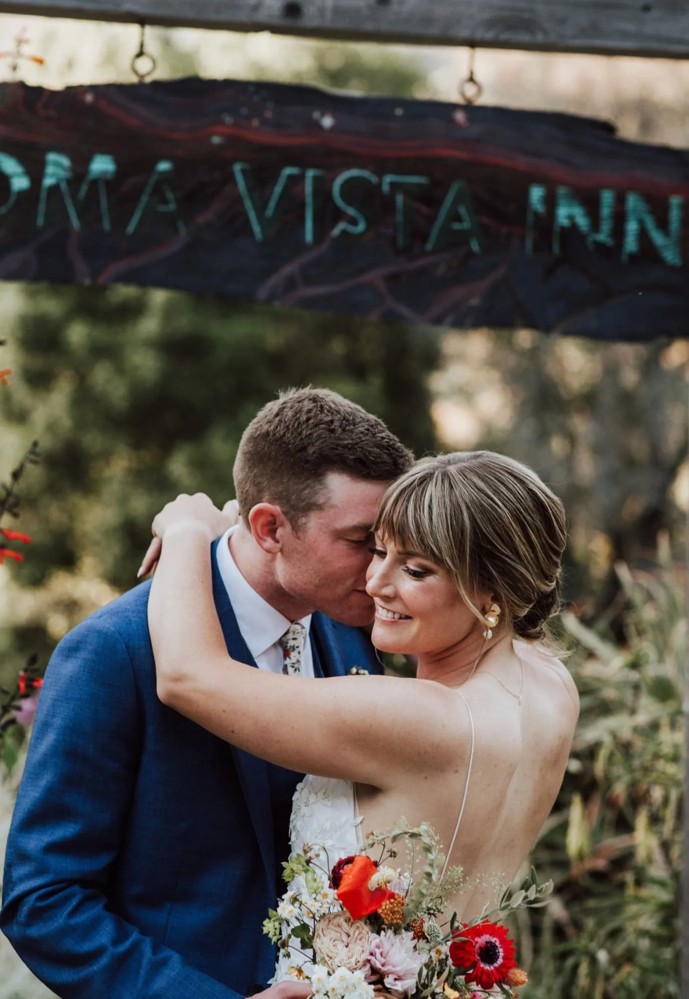A newlywed couple sharing a dance outdoors, with the bride holding a bouquet of colorful flowers, and a sign in the background reading 'ON A VISTA INN'.