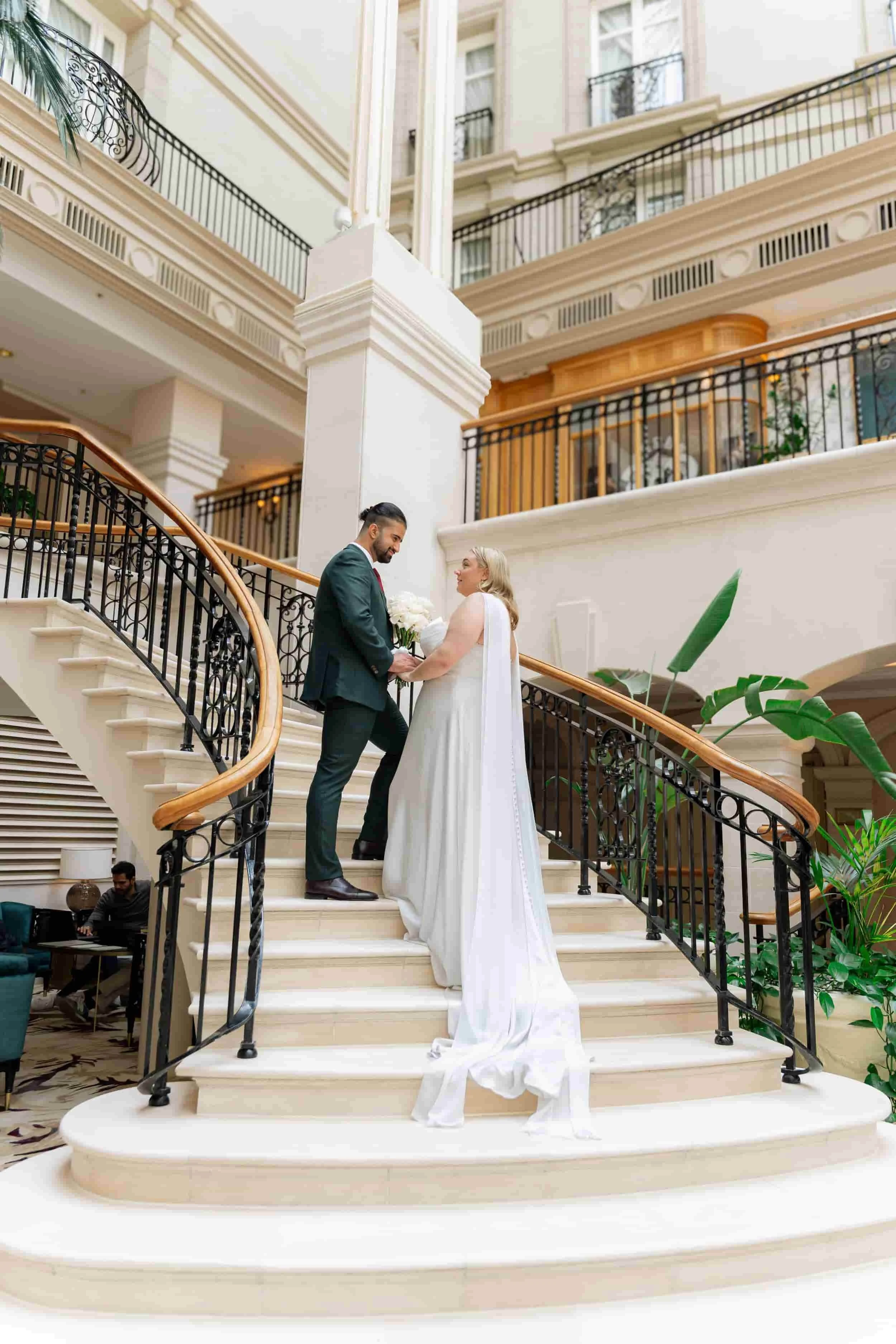 A bride and groom standing on a grand staircase inside a luxurious hotel, holding hands and smiling at each other during their wedding ceremony.