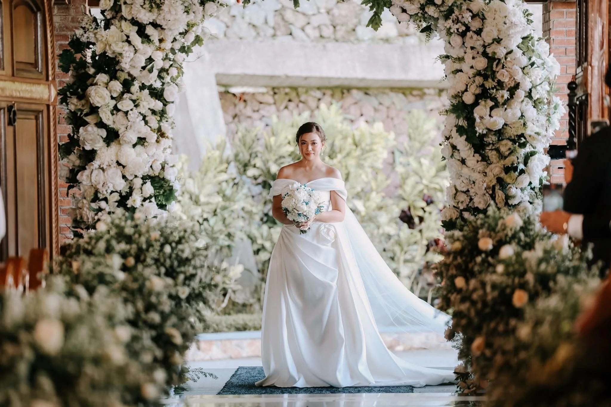 A bride in a white off-shoulder wedding gown holding a bouquet walking through a floral archway decorated with white roses and greenery at an outdoor wedding venue.