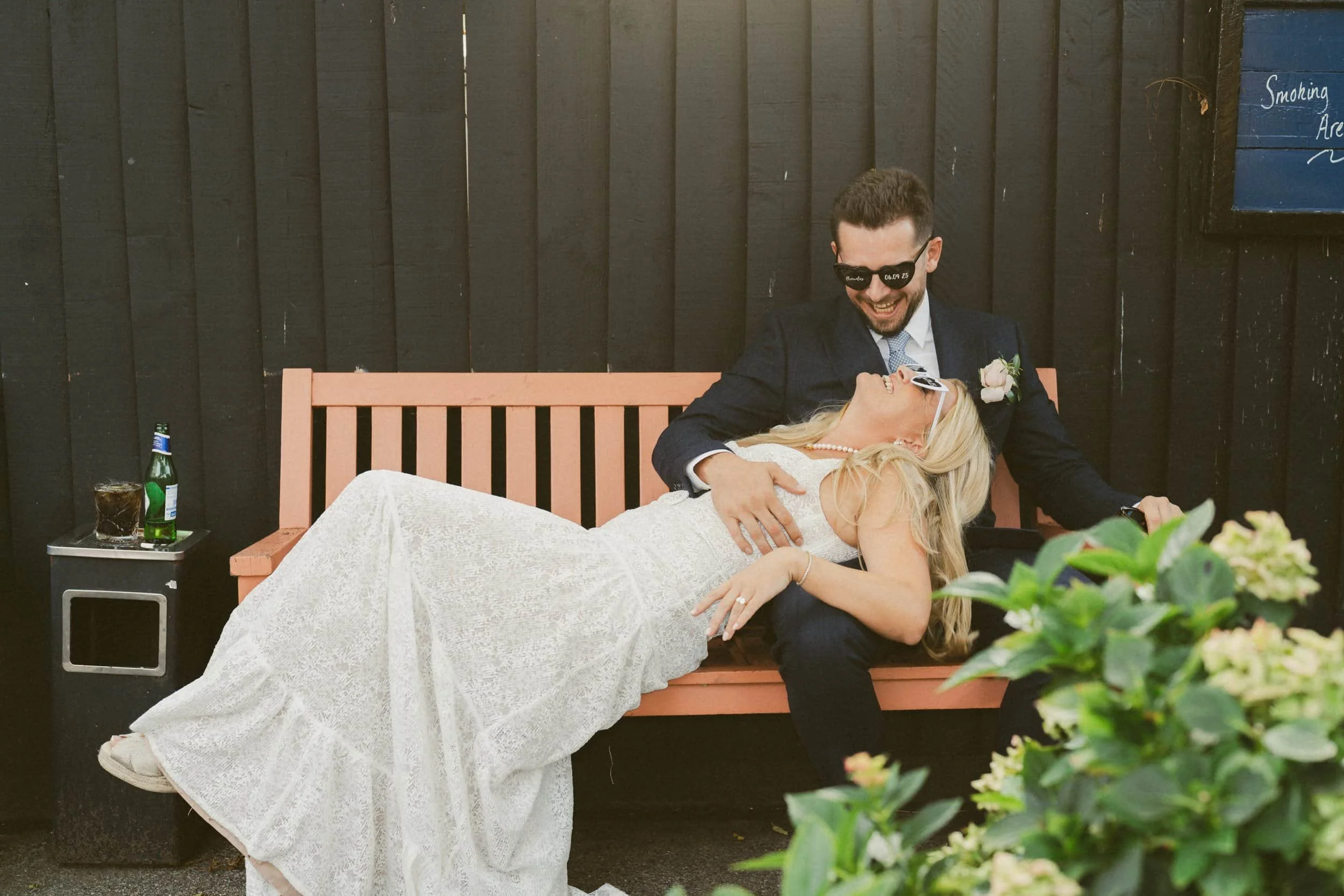A newlywed couple celebrating outdoors, sitting on a pink wooden bench, with the groom in a dark suit and sunglasses, holding the bride, who is in a white lace wedding dress, lying on his lap. There are drinks and a small table beside them, and a dar