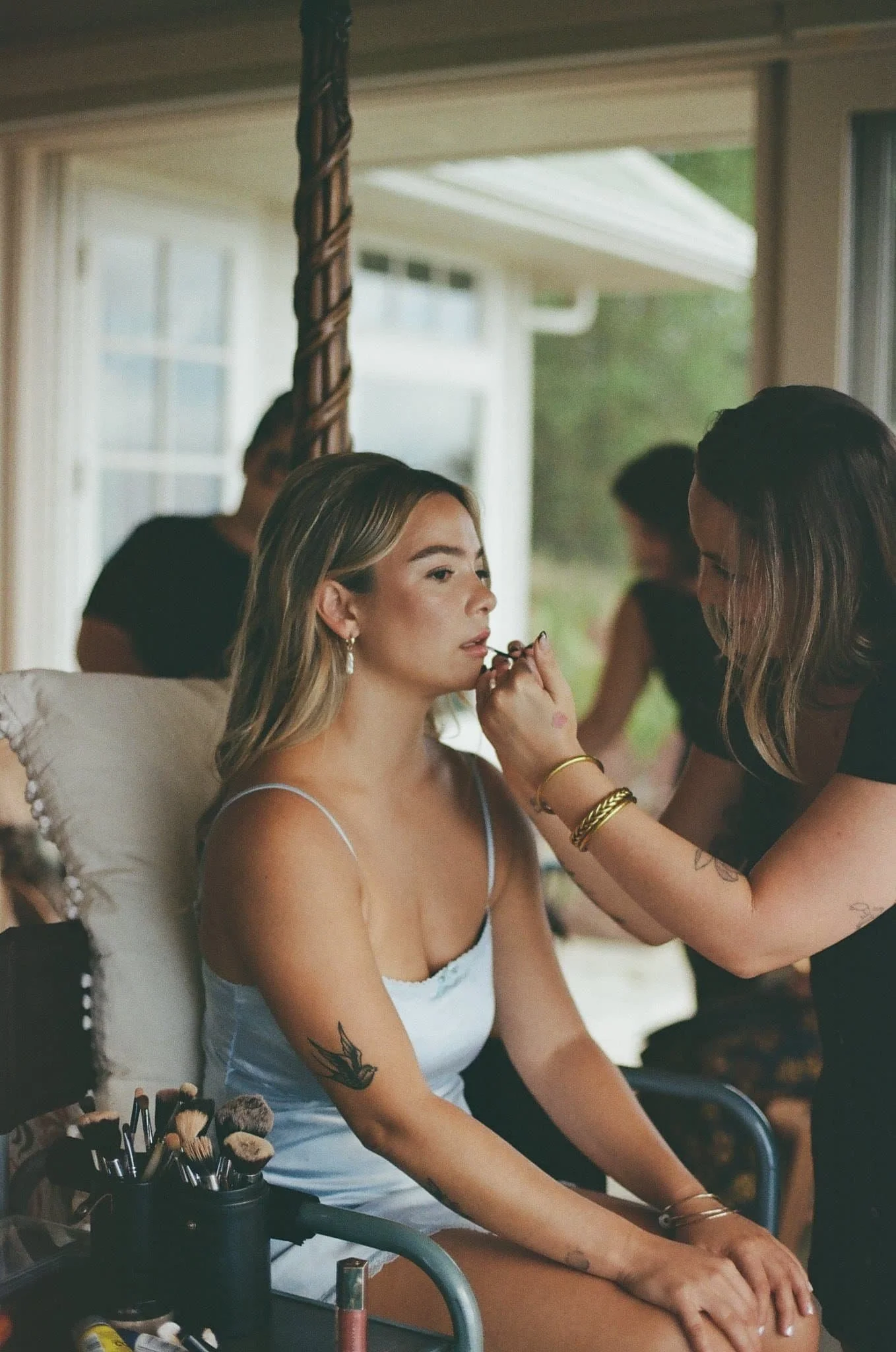 A woman with blonde hair and a tattoo of a bird on her right arm sitting outdoors in front of a window, having her makeup applied by a makeup artist. Makeup brushes and beauty products are on a nearby table.