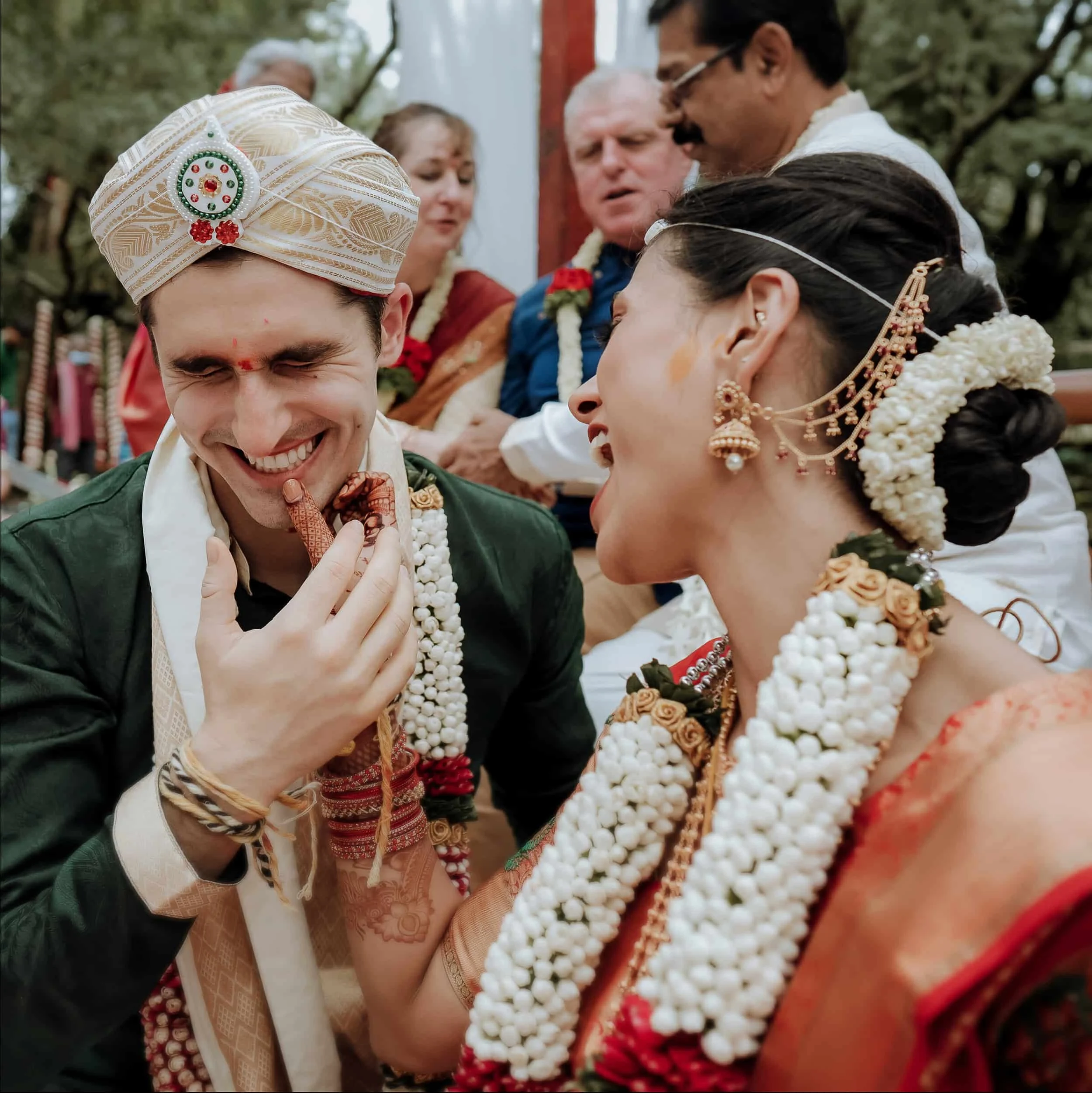 Indian couple dressed in traditional attire during a wedding ceremony, exchanging flower garlands surrounded by family members.