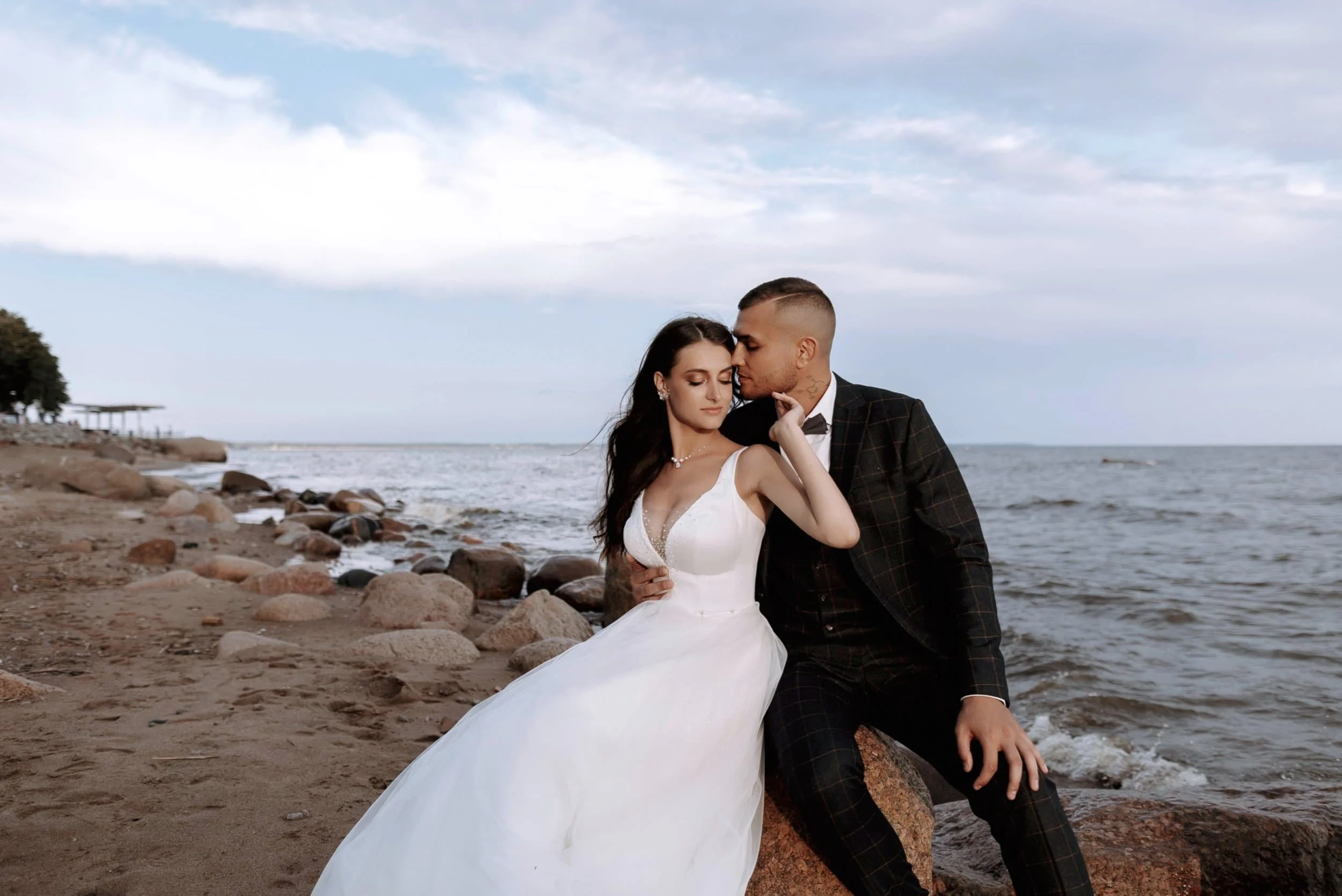 A couple dressed in wedding attire embracing on a rocky beach with the ocean and cloudy sky in the background.