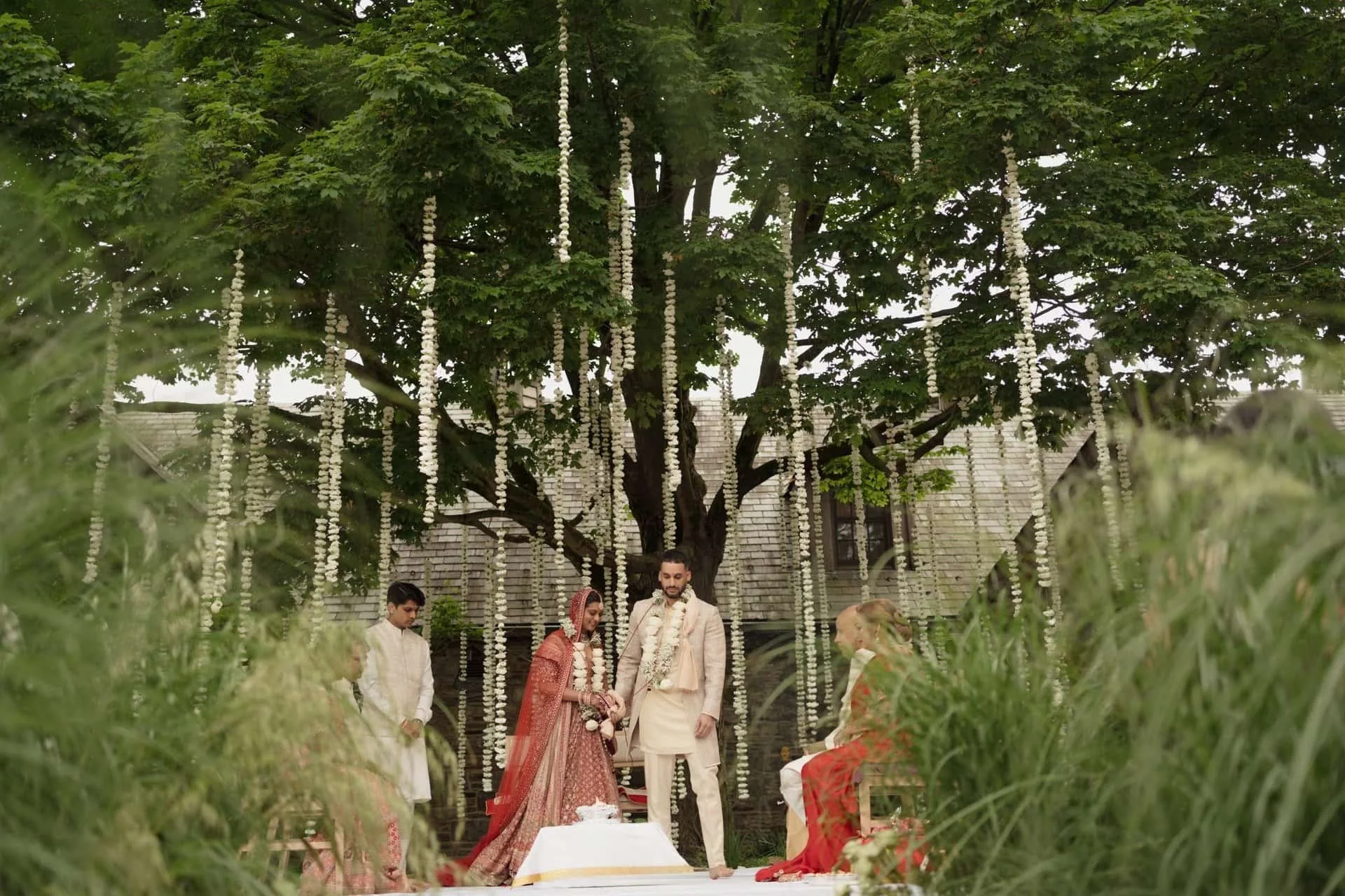 A wedding ceremony taking place outdoors beneath a large green tree decorated with hanging white flower garlands. A bride in traditional red and gold attire is standing with the groom, surrounded by friends and family, with some seated on the grass.