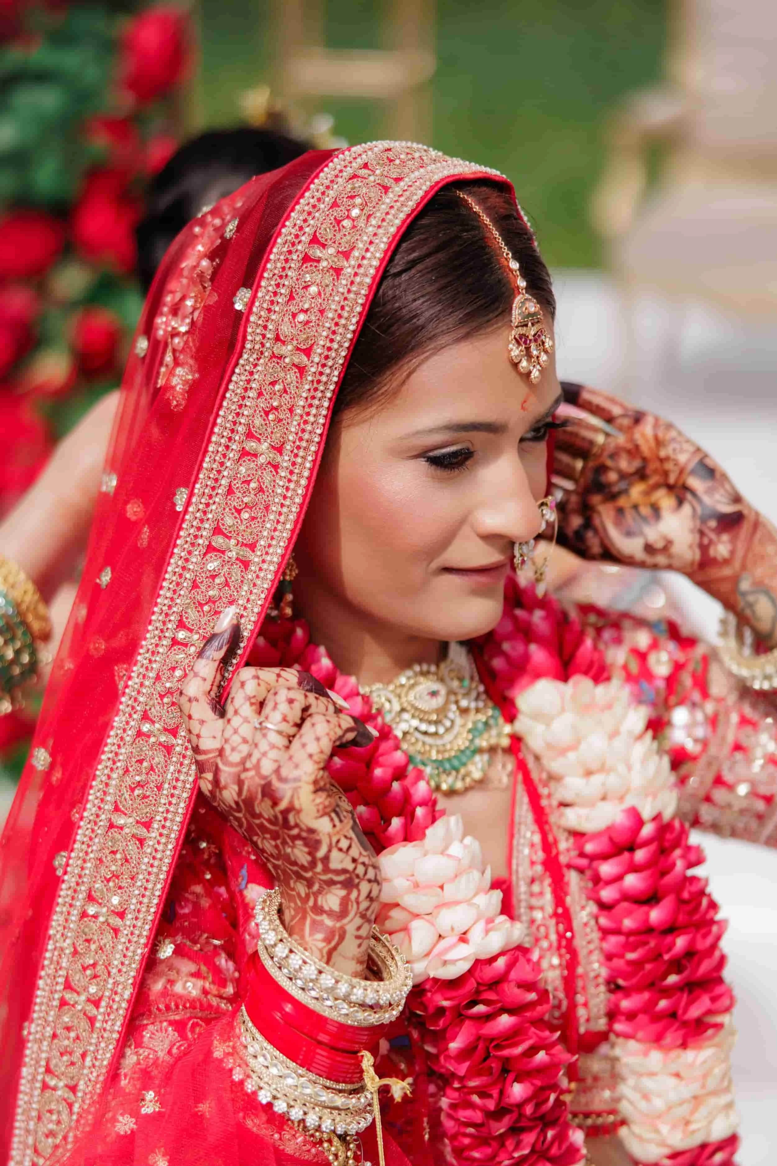 Close-up of a woman in traditional Indian bridal attire, including a red and gold saree with intricate embroidery, jewelry with pearls and gemstones, and a floral garland.