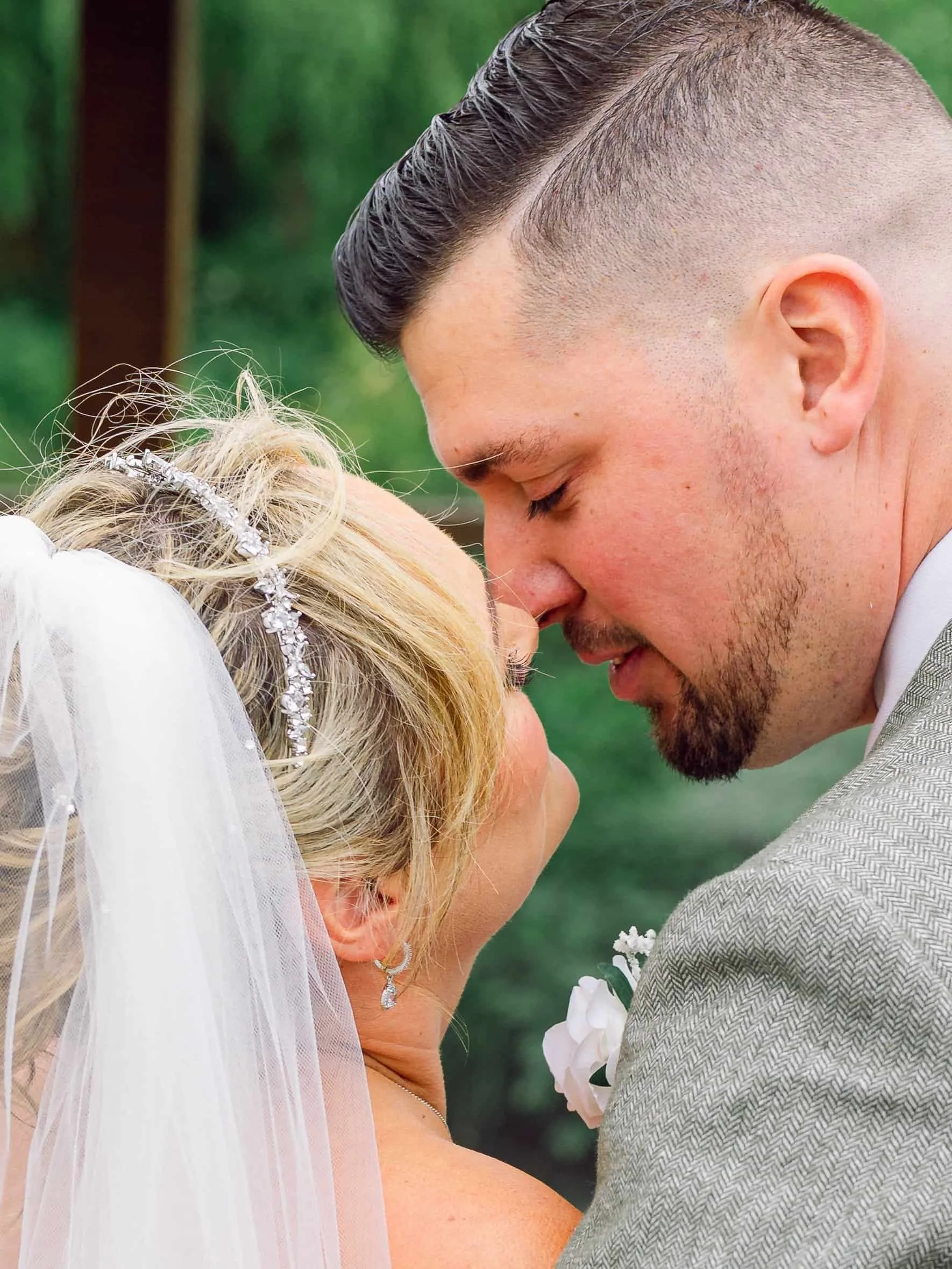 A close-up of a couple during their wedding, with their foreheads touching and eyes closed, outdoors with a green background.