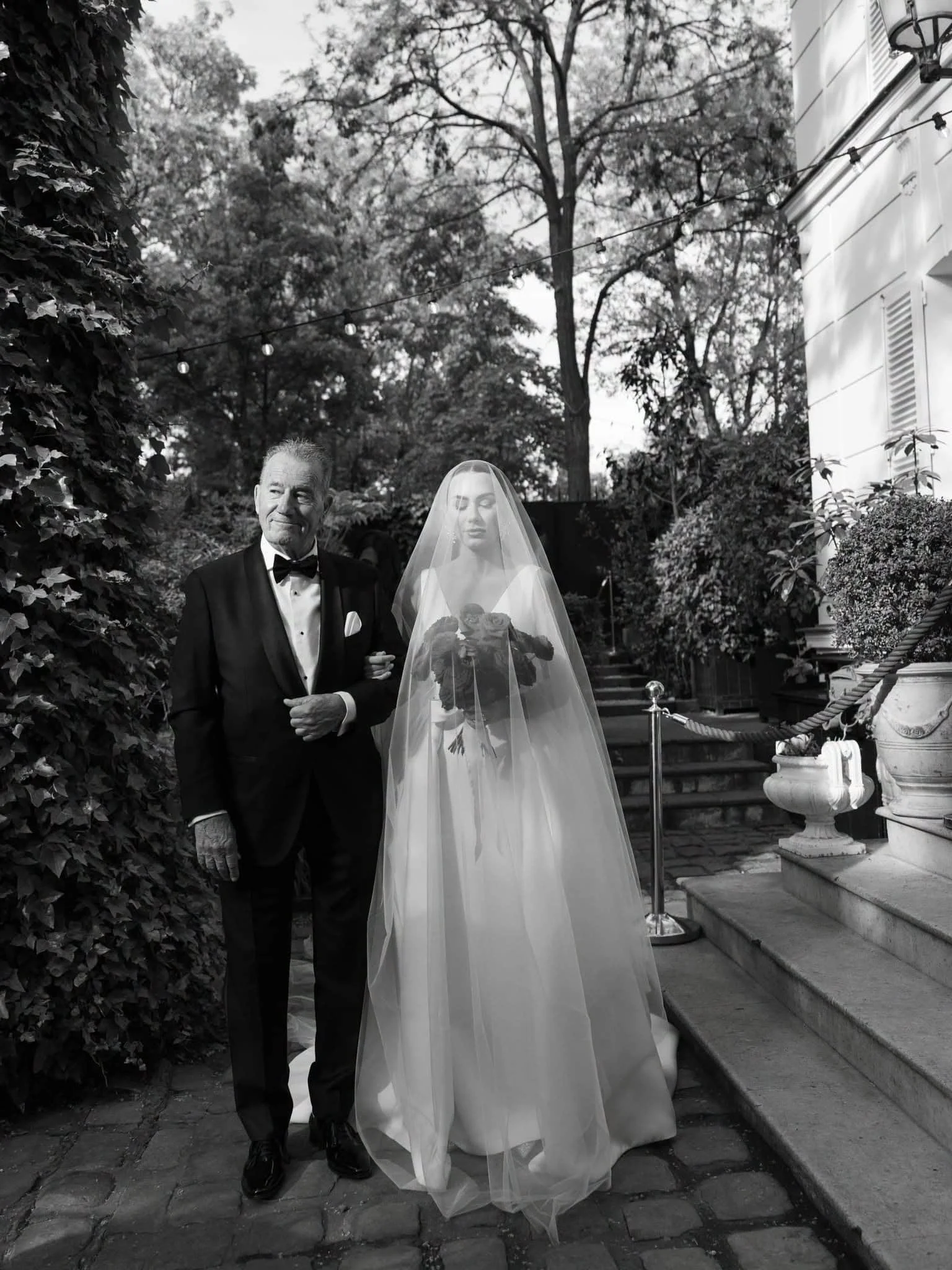A man in a tuxedo walking with a woman in a wedding dress and veil, holding a bouquet of roses, outdoors on a stone path near stairs, with trees, bushes, and a building in the background.