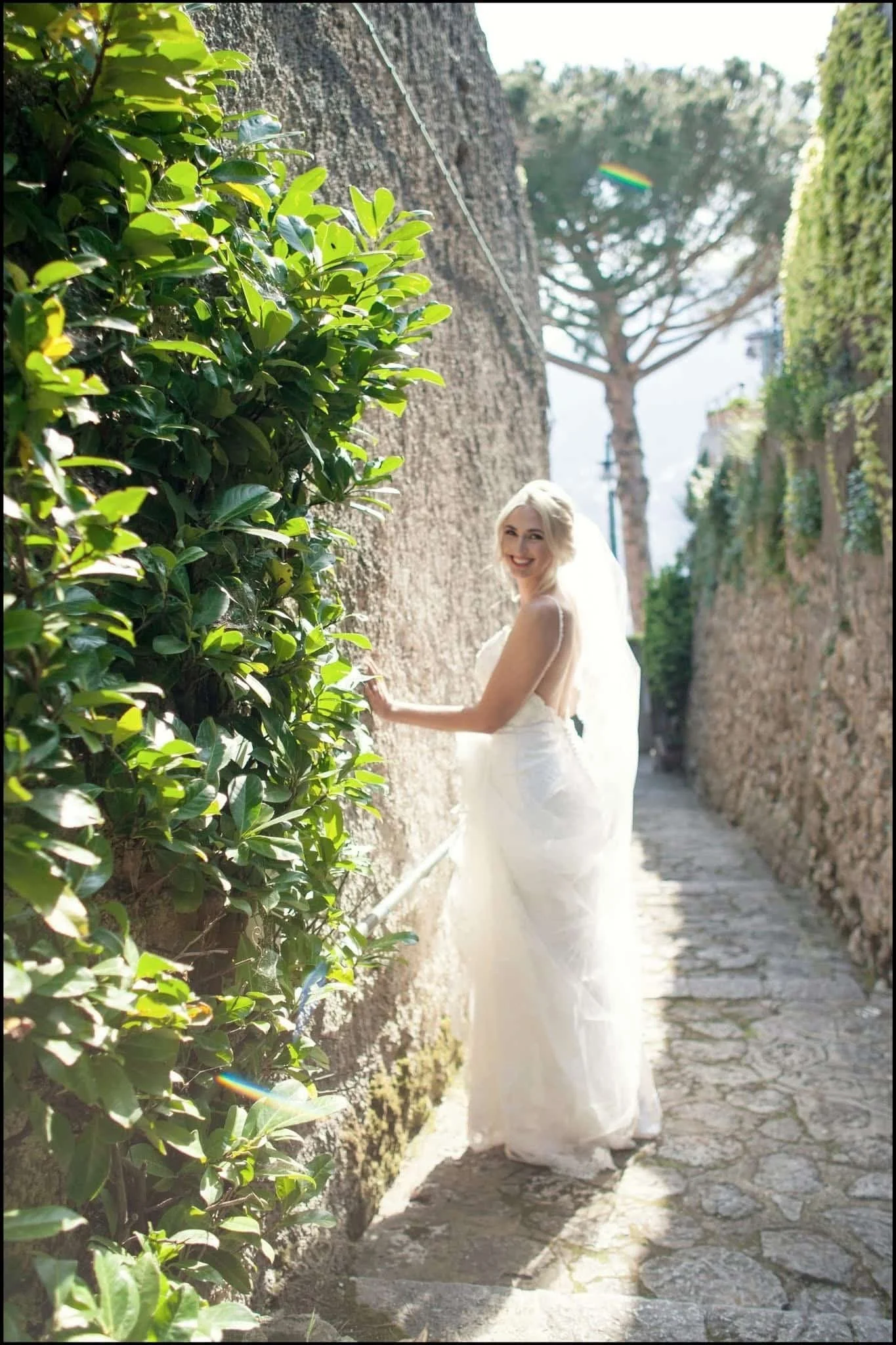 A smiling woman in a white wedding dress and veil standing in a narrow alley with a tall brick wall on one side covered with green bushes and a large tree in the background with a rainbow-colored prism effect in the sky.