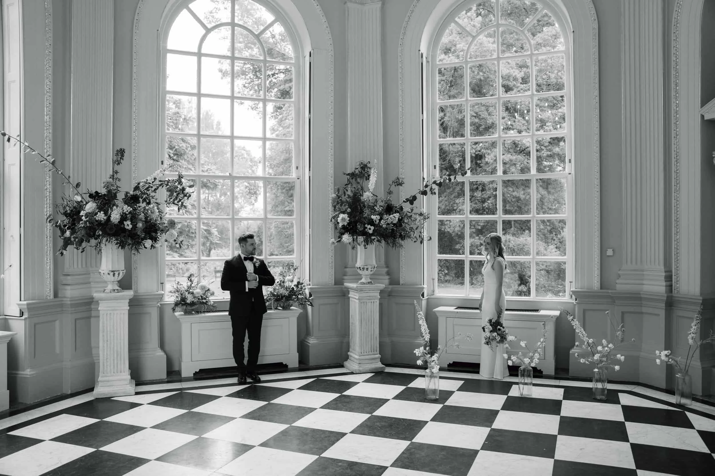 A bride and groom standing in a grand, ornate room with large arched windows, decorative columns, and floral arrangements for a wedding ceremony.