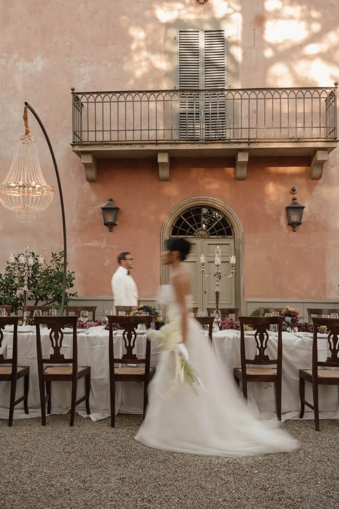 Indoor wedding reception with a long table covered in white tablecloths, set for guests, and a bride in a white gown walking past. There are wooden chairs, candelabras, and floral decorations on the table. The background features a pinkish wall with 