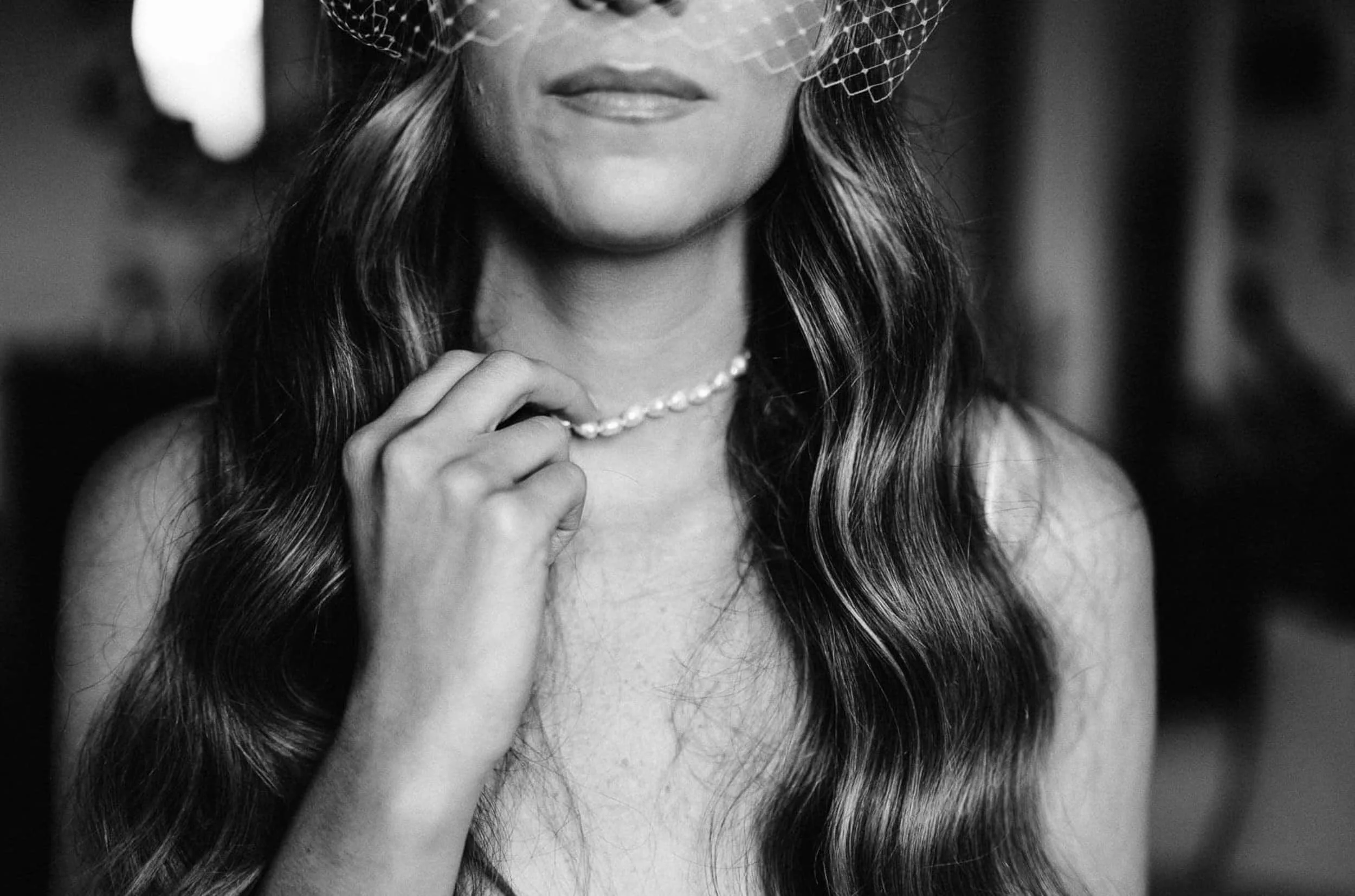 A black and white close-up photo of a woman wearing a fishnet veil over her eyes, holding a pearl necklace with her hand, with long wavy hair and a slight smile.