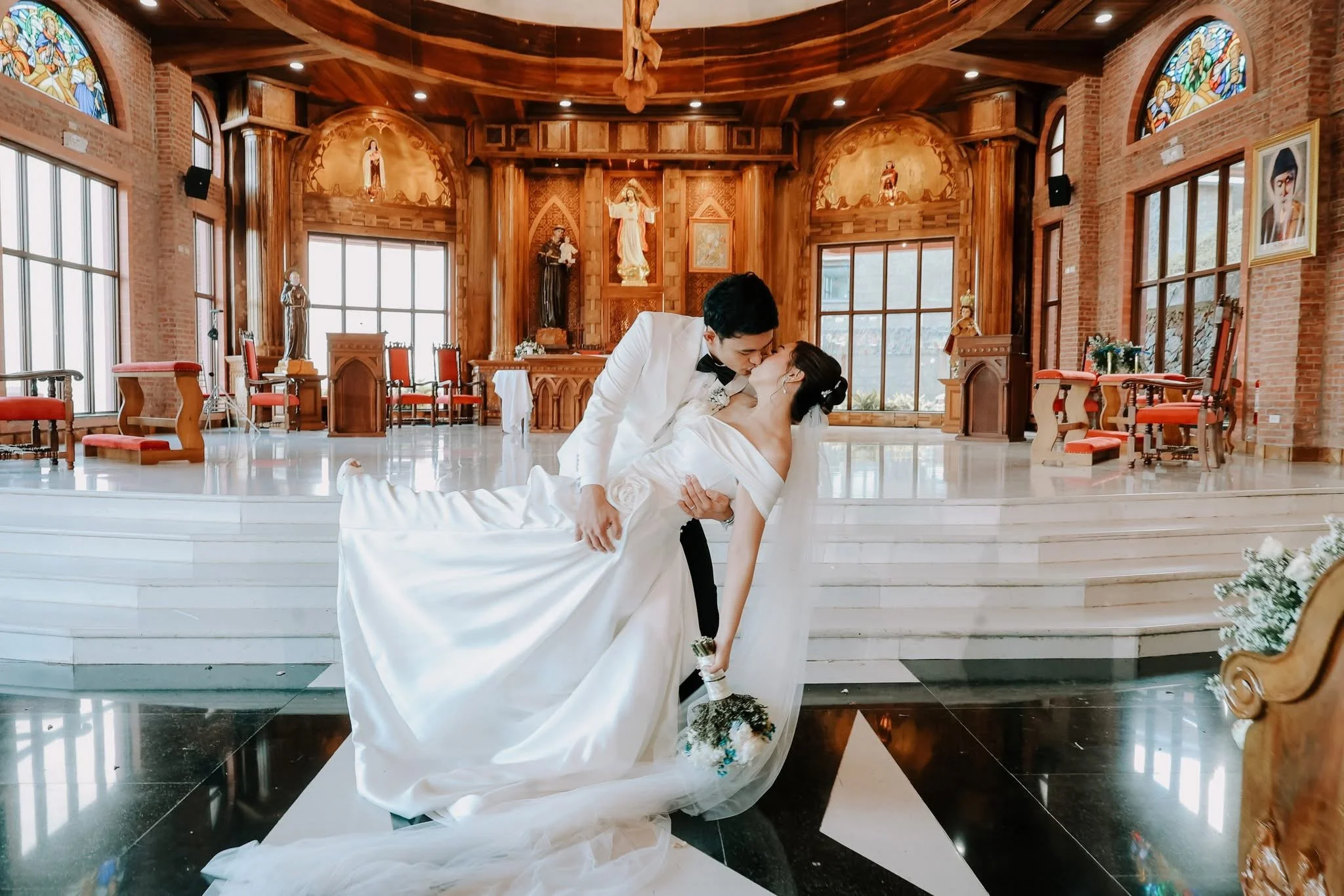 A newlywed couple is dancing and kissing inside a church with wooden decor, stained glass windows, and religious statues. The bride is in a white wedding dress and the groom is in a tuxedo.