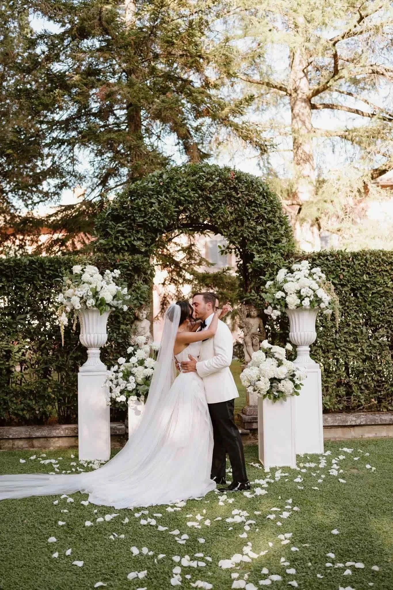 A bride and groom sharing a kiss at their wedding ceremony outdoors, surrounded by white floral arrangements on pillars and a lush green garden with trees in the background.