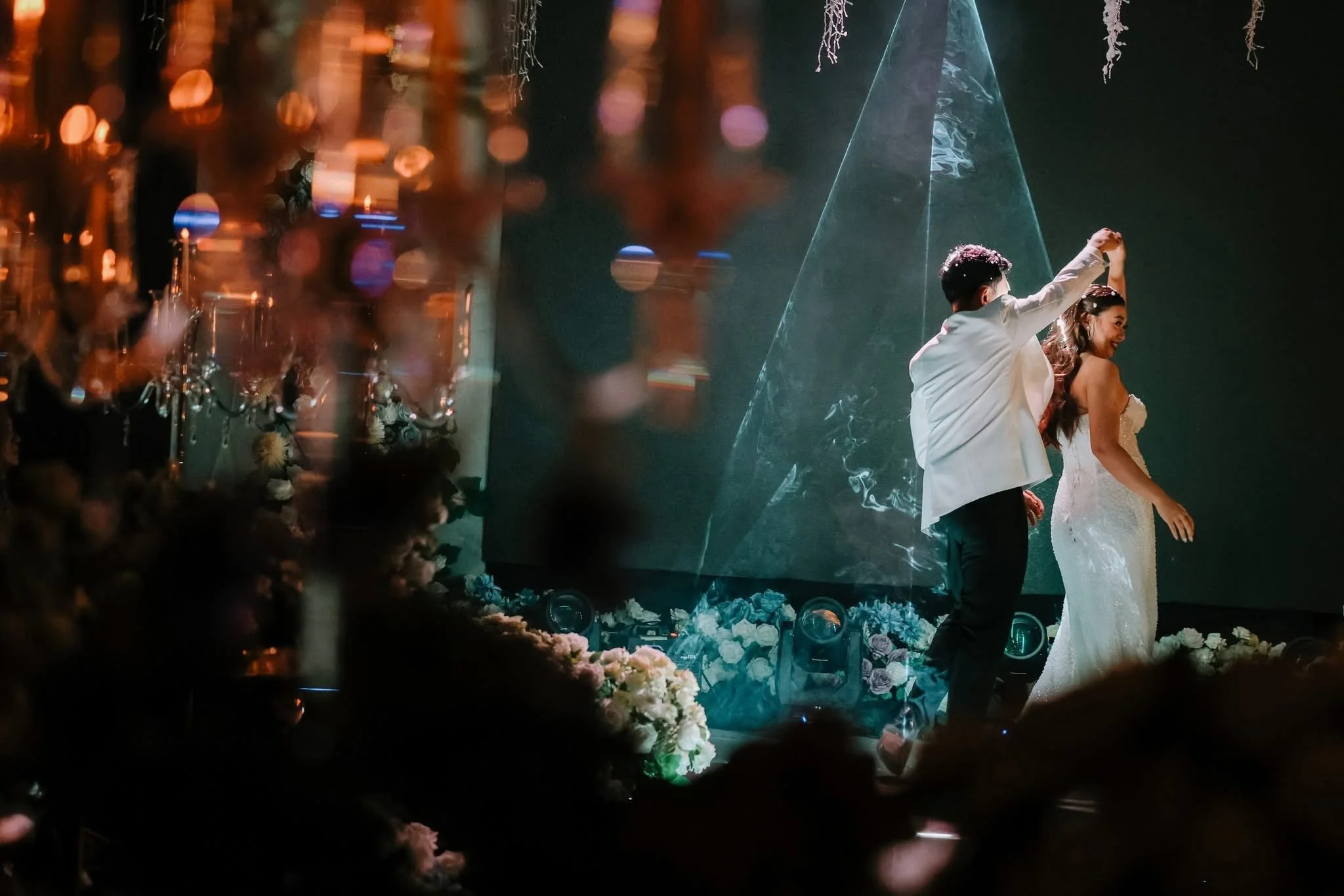 A newlywed couple dancing at their wedding reception, with floral decorations and a dark backdrop.