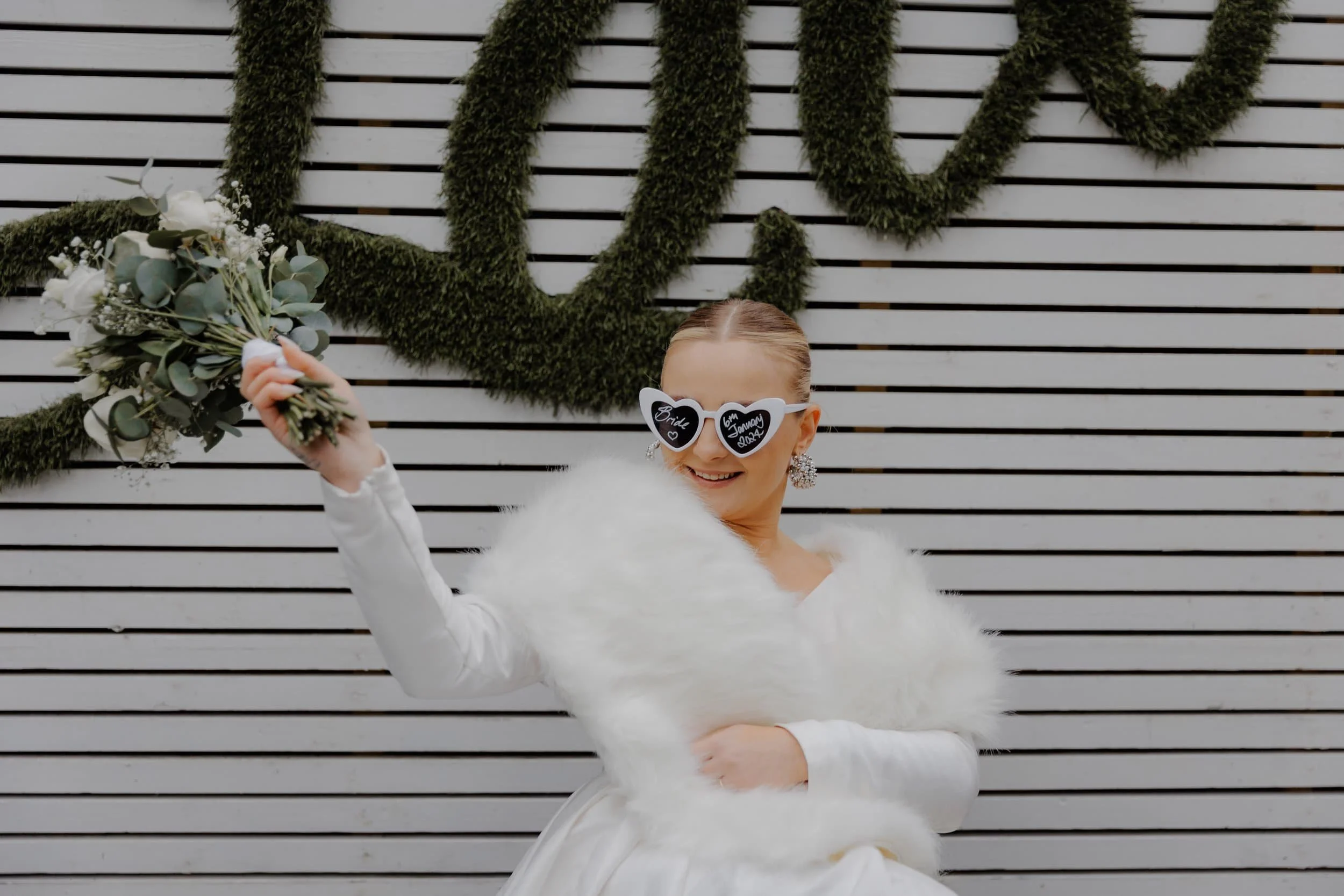 A woman in a white dress and fluffy white shawl, wearing heart-shaped sunglasses with messages 'Bride' and 'Groom' on each lens, is holding a bouquet of white flowers and greenery. She is standing in front of a white wooden fence with moss-covered le
