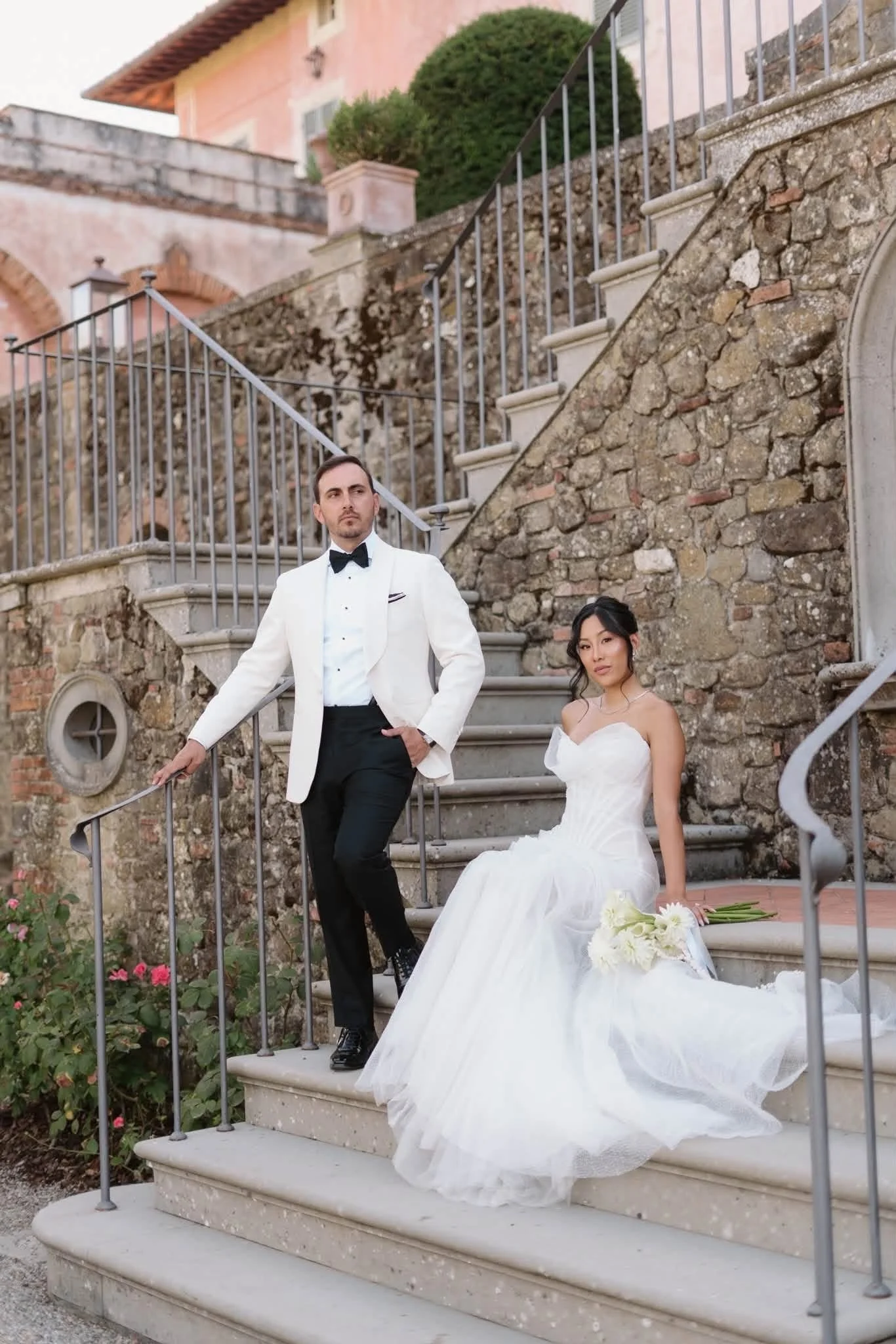 A man in a white tuxedo jacket and black pants stands with a woman in a white wedding dress, sitting on stone steps outside a building with a stone wall and green shrubs in the background. The woman holds a bouquet of white flowers, and a bouquet lie