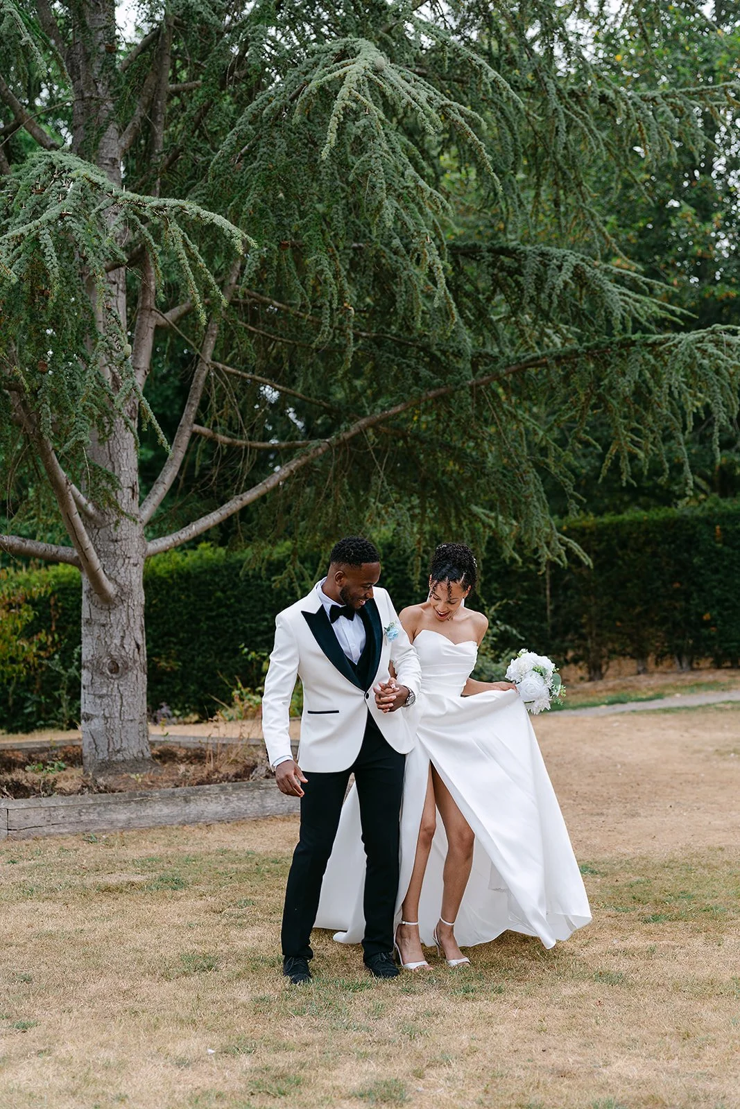 A bride and groom smiling and holding hands outdoors under a large tree, the bride in a white wedding gown with a slit, and the groom in a white tuxedo jacket with black lapel and black pants.