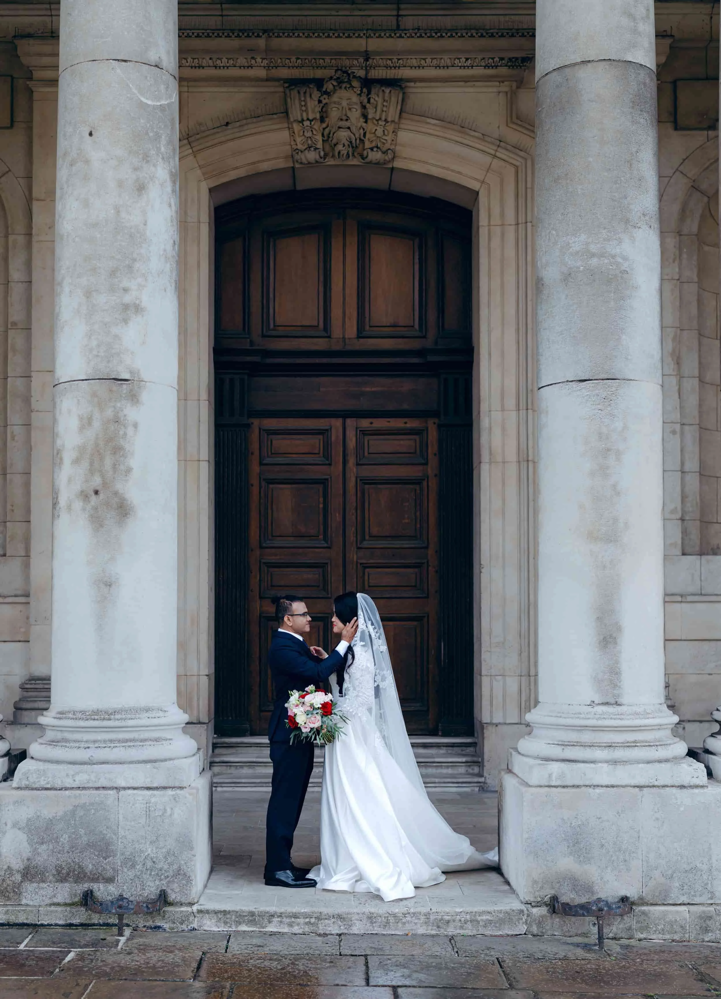 A bride and groom standing close together in front of a large wooden door, framed by two tall stone columns on a church exterior. The groom wears a suit and holds a bouquet, while the bride wears a white wedding gown and veil.