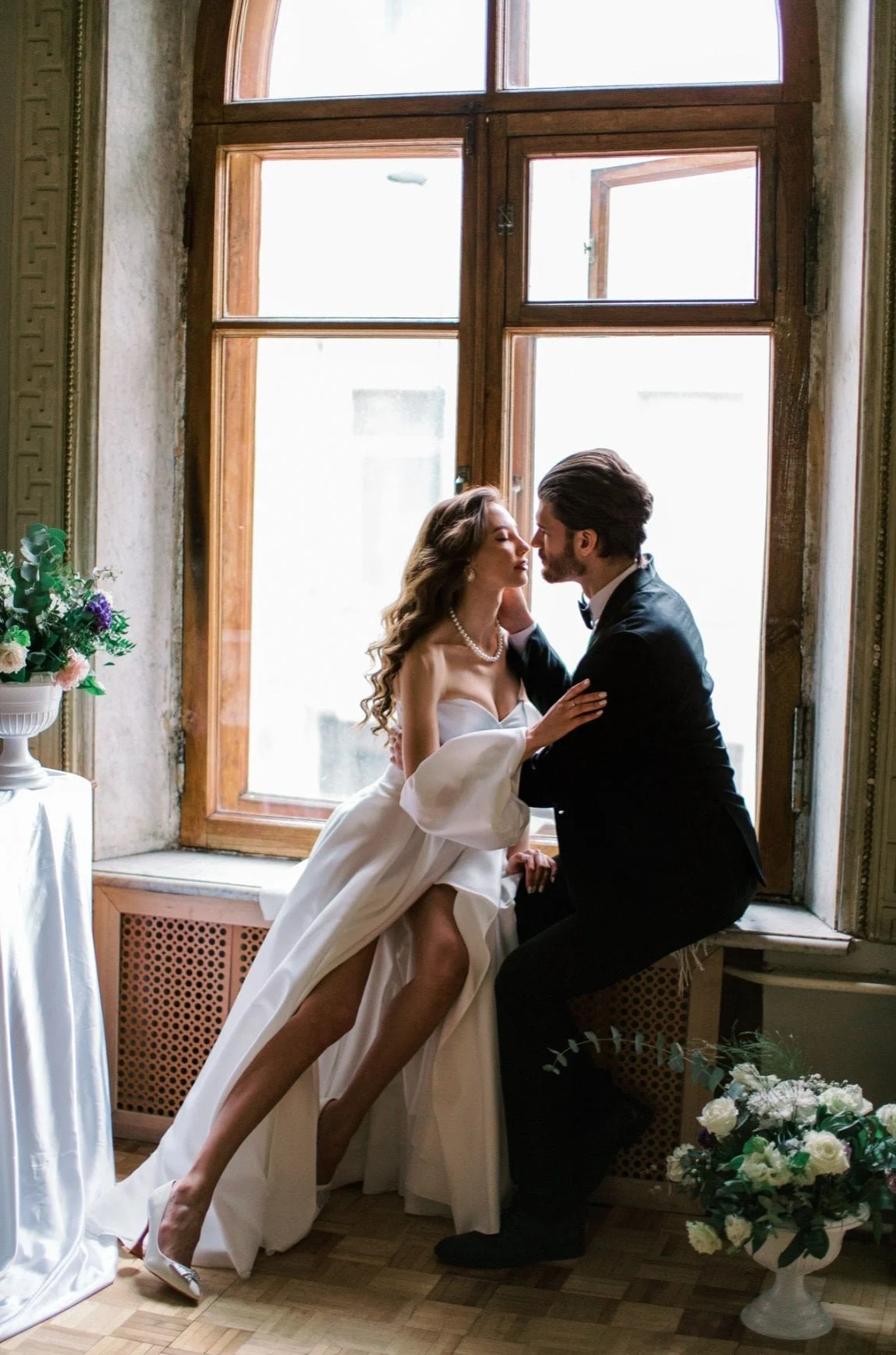 A bride and groom kiss in front of a large window, with flowers nearby, during a wedding ceremony.