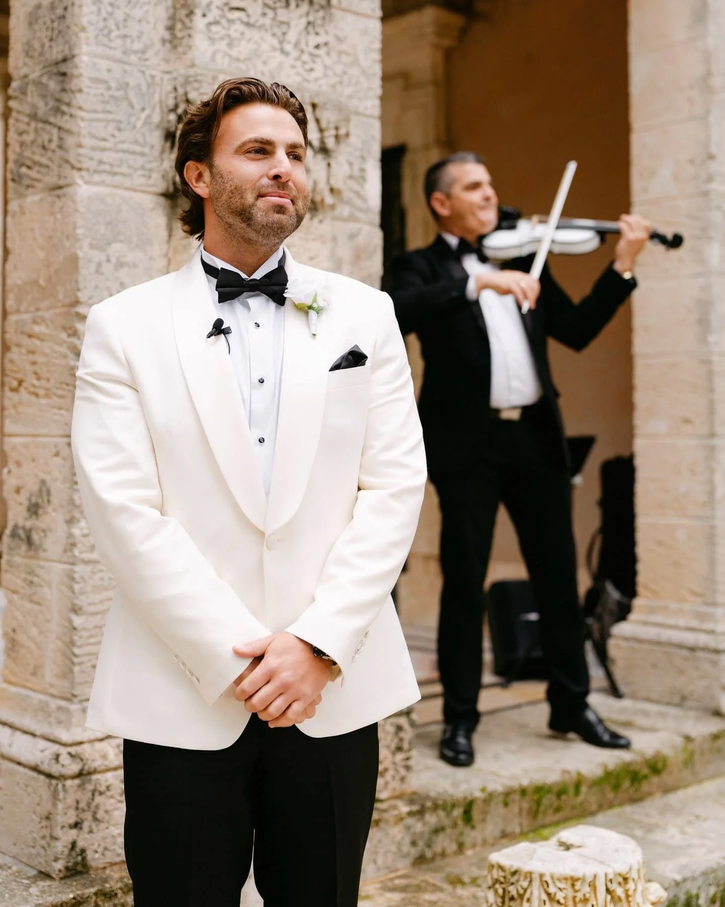 A groom in a white tuxedo with a black bow tie standing with his hands clasped together, in front of ancient stone ruins, with a violinist in a black tuxedo playing in the background.