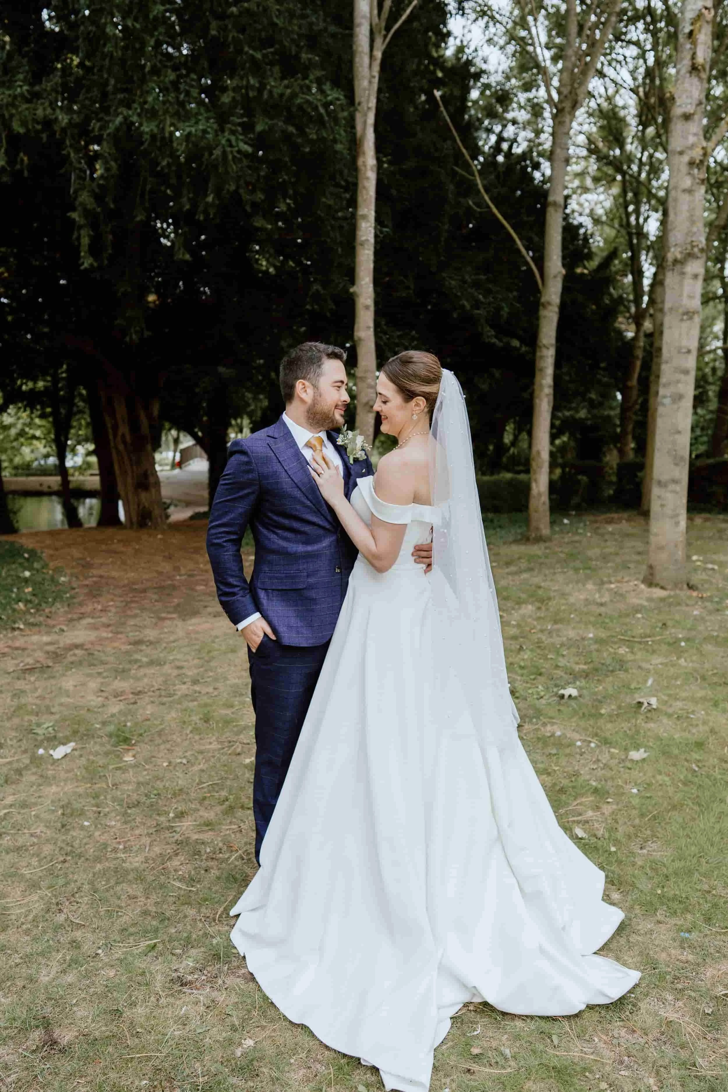 A bride and groom standing close together outdoors in a park, smiling and looking into each other's eyes. The bride is wearing an off-the-shoulder white wedding gown and a veil, while the groom is dressed in a blue checkered suit with a tie. Tall tre