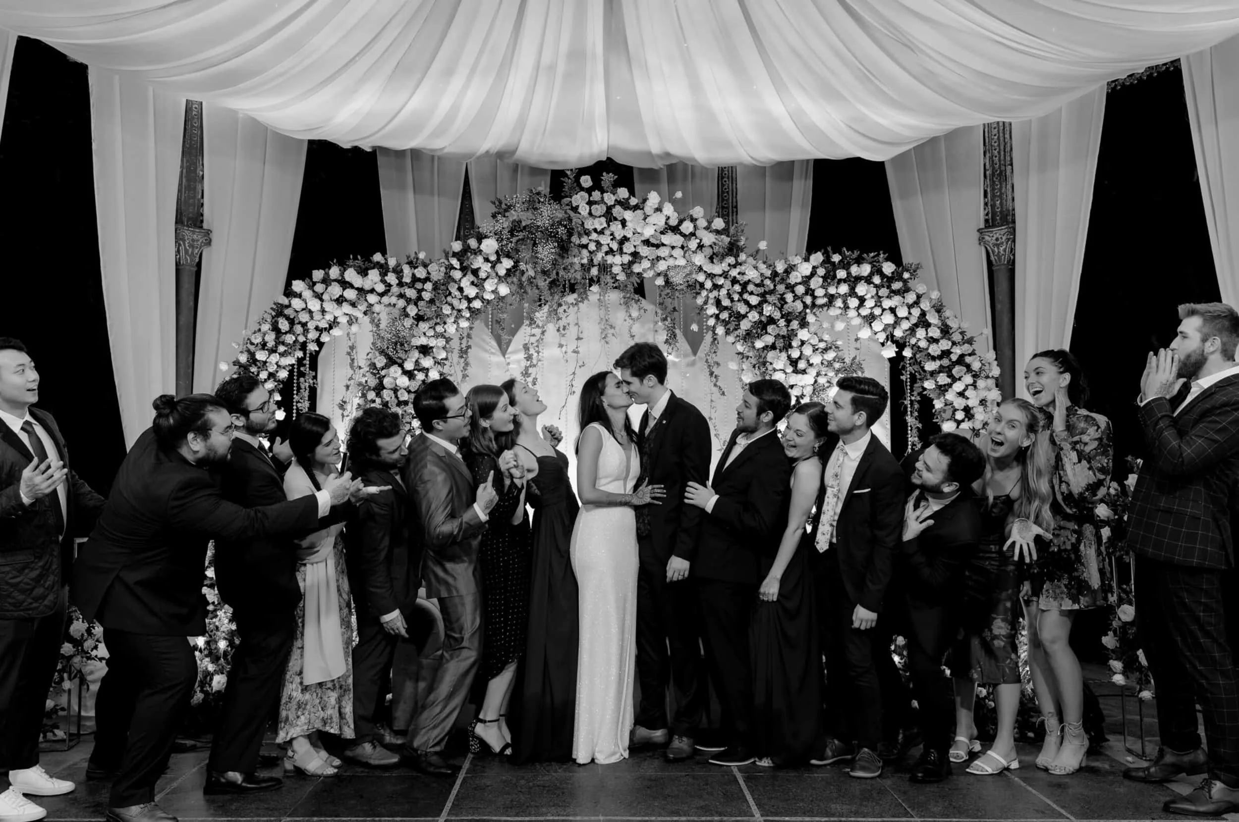 A black-and-white photo of a wedding reception with the bride and groom sharing a kiss in front of an arch decorated with flowers, surrounded by guests celebrating and taking photos.