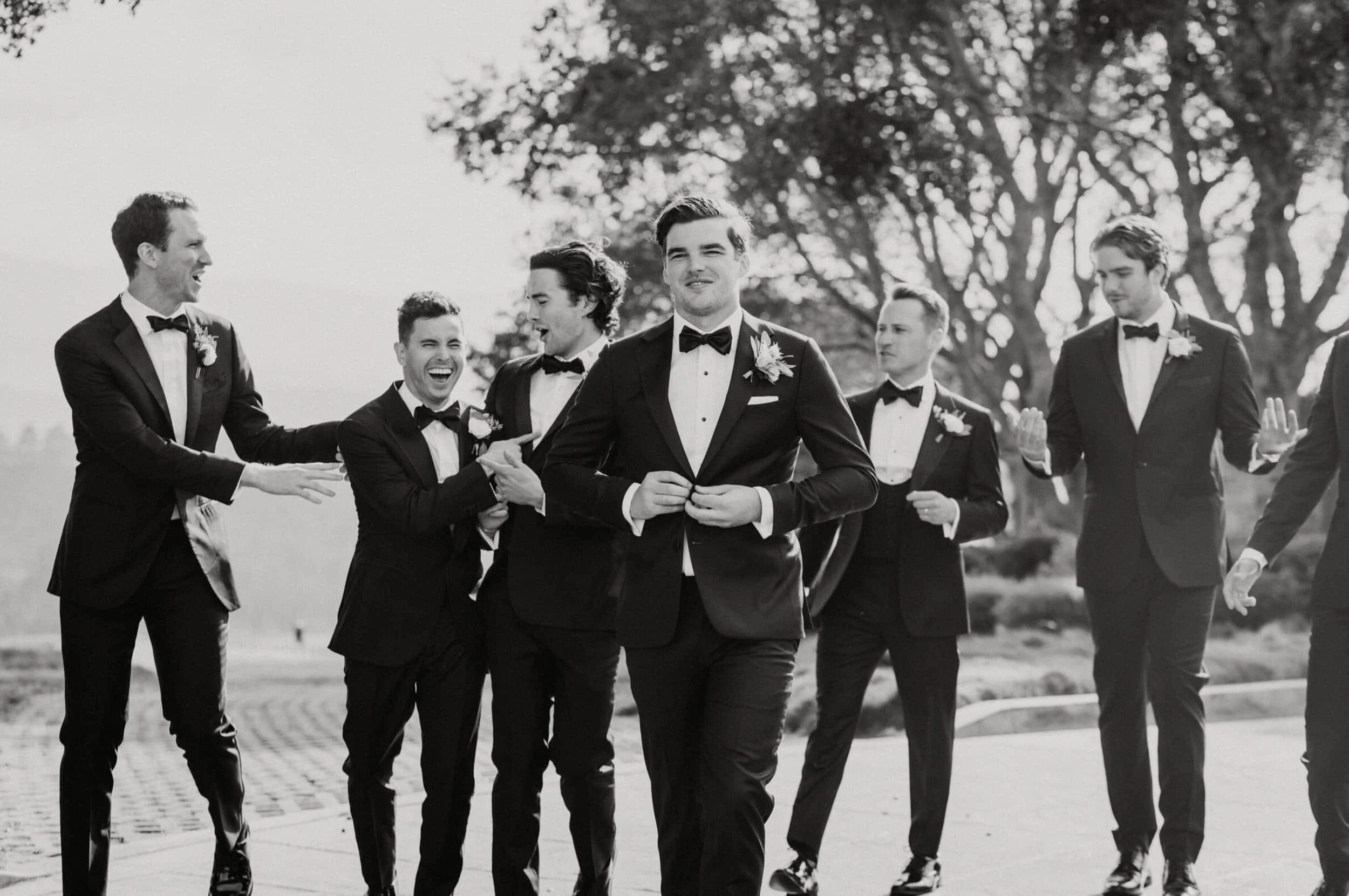Group of men dressed in tuxedos with bow ties, walking outdoors in a park, smiling and engaging with each other, with large trees in the background.