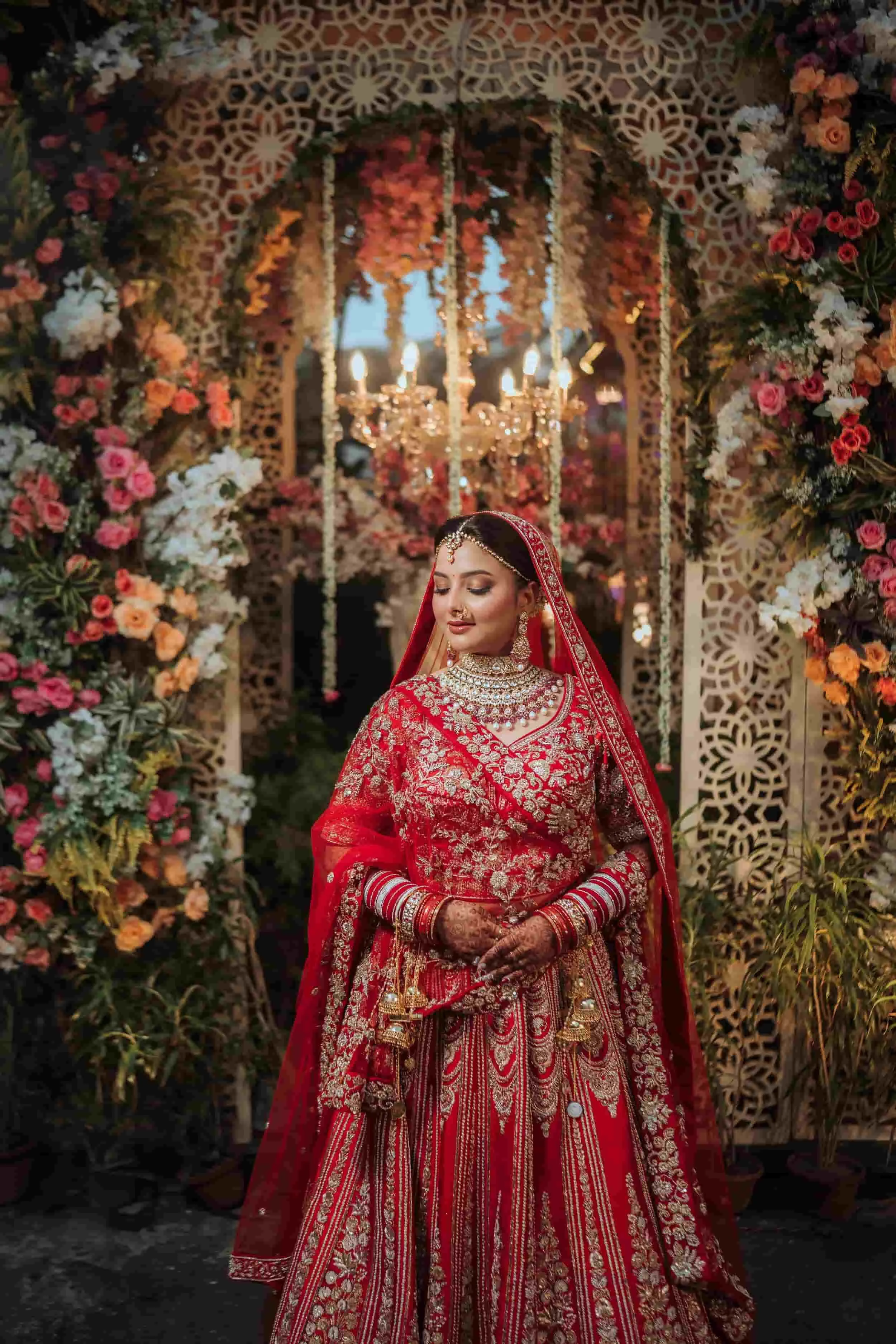 A woman dressed in a red and gold traditional wedding outfit, standing in front of floral and decorative backdrop with chandeliers and ornate patterns.