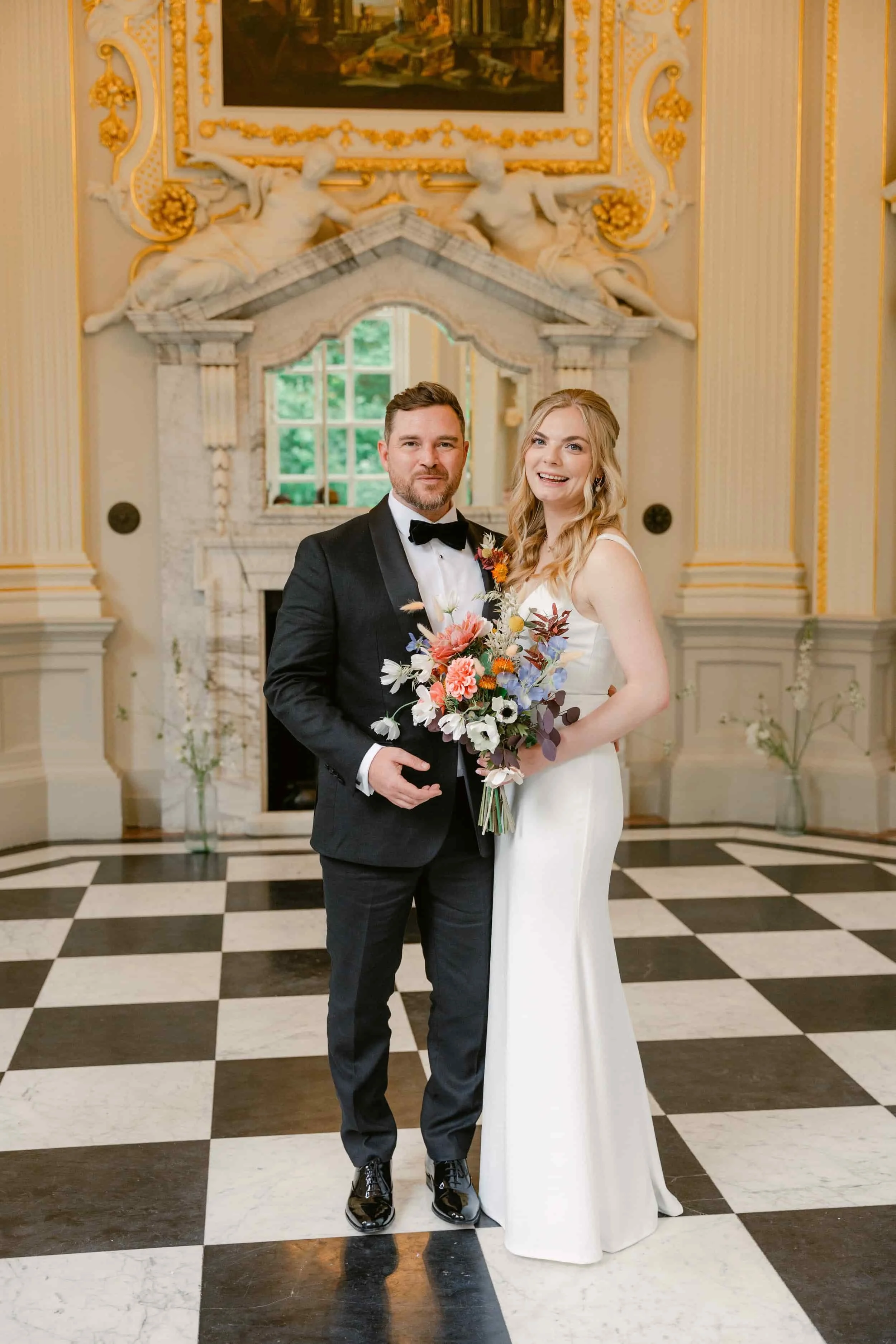 A newlywed couple standing together in an ornate room. The groom is wearing a black tuxedo with a bow tie, and the bride is in a white wedding dress holding a bouquet of colorful flowers. The background features a decorative marble fireplace, large w
