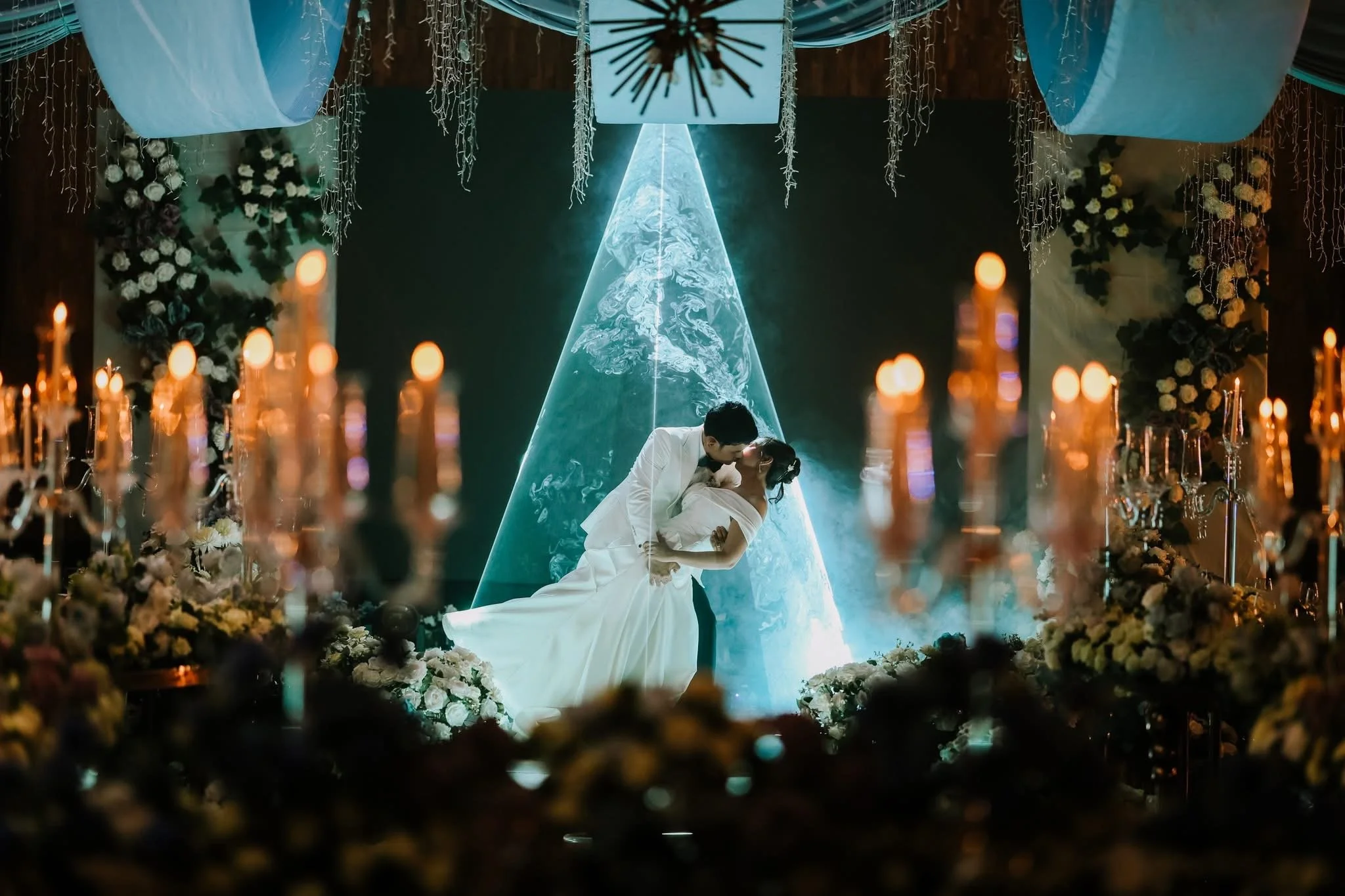 A bride and groom sharing a kiss during their wedding ceremony, with the bride in a white gown and the groom in a white suit, surrounded by floral arrangements and candles, on a stage with a digital globe and winged structures in the background.