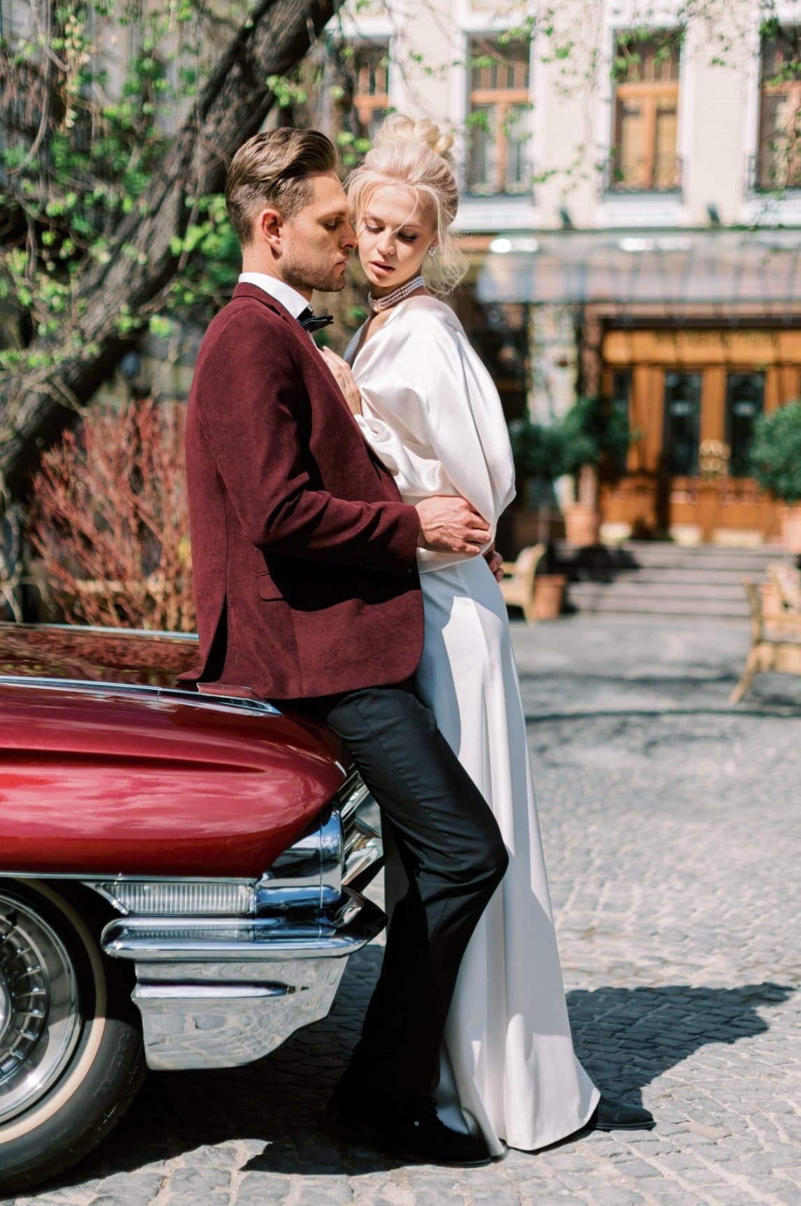 A man in a maroon tuxedo jacket and a woman in a white dress leaning against a vintage car outdoors in front of a building with trees and windows.