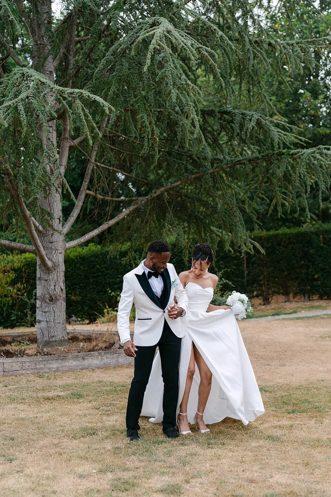 A newlywed couple walking outdoors under a large tree, with the groom in a white tuxedo jacket and the bride in a strapless white wedding gown, holding a bouquet, smiling and enjoying their moment.