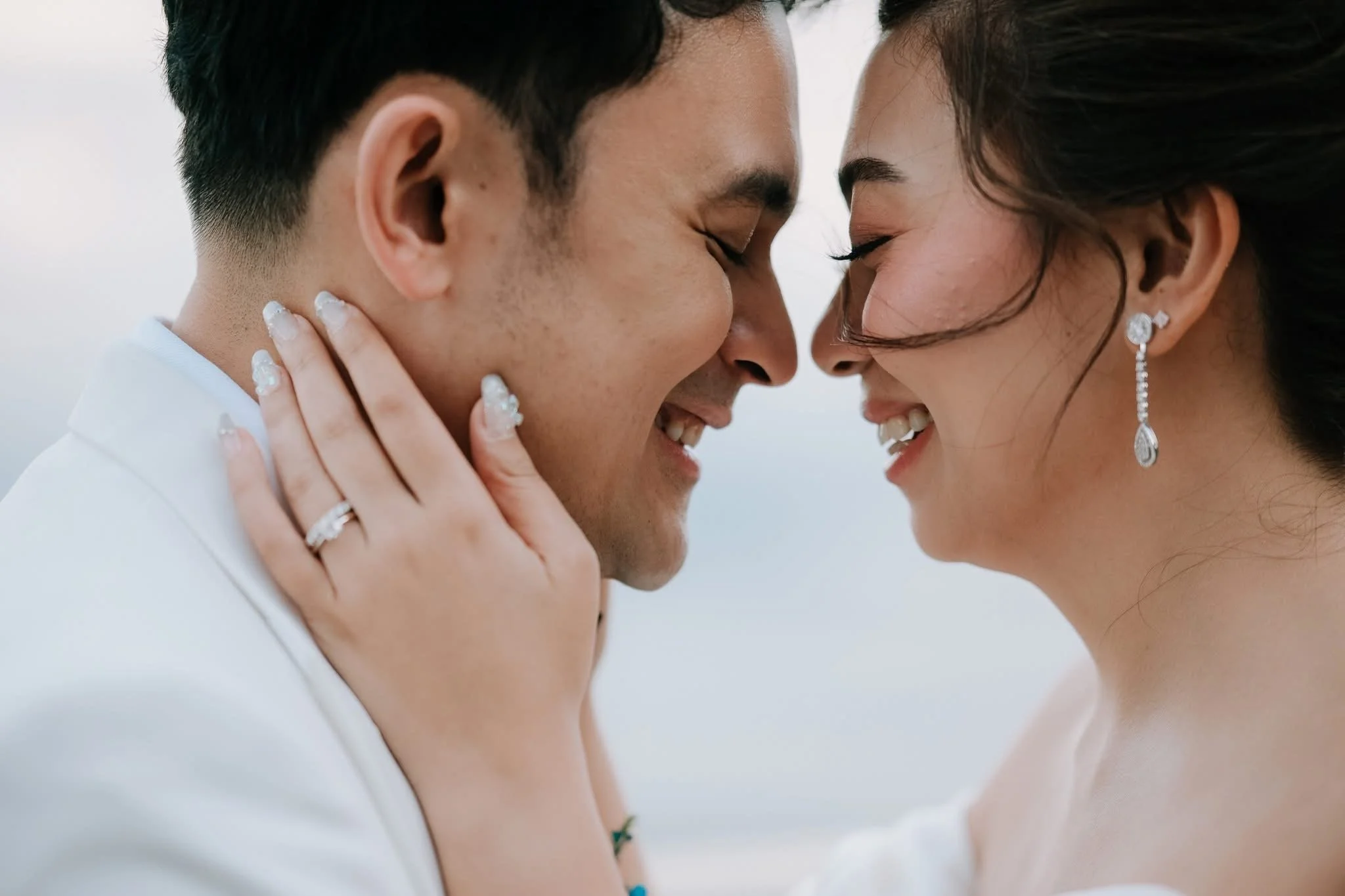 A close-up of a couple with foreheads touching, smiling, with eyes closed. The woman has dark hair, diamond earrings, and jewelry, while the man has short dark hair and a white shirt. Their faces are very close, and they appear happy and intimate.