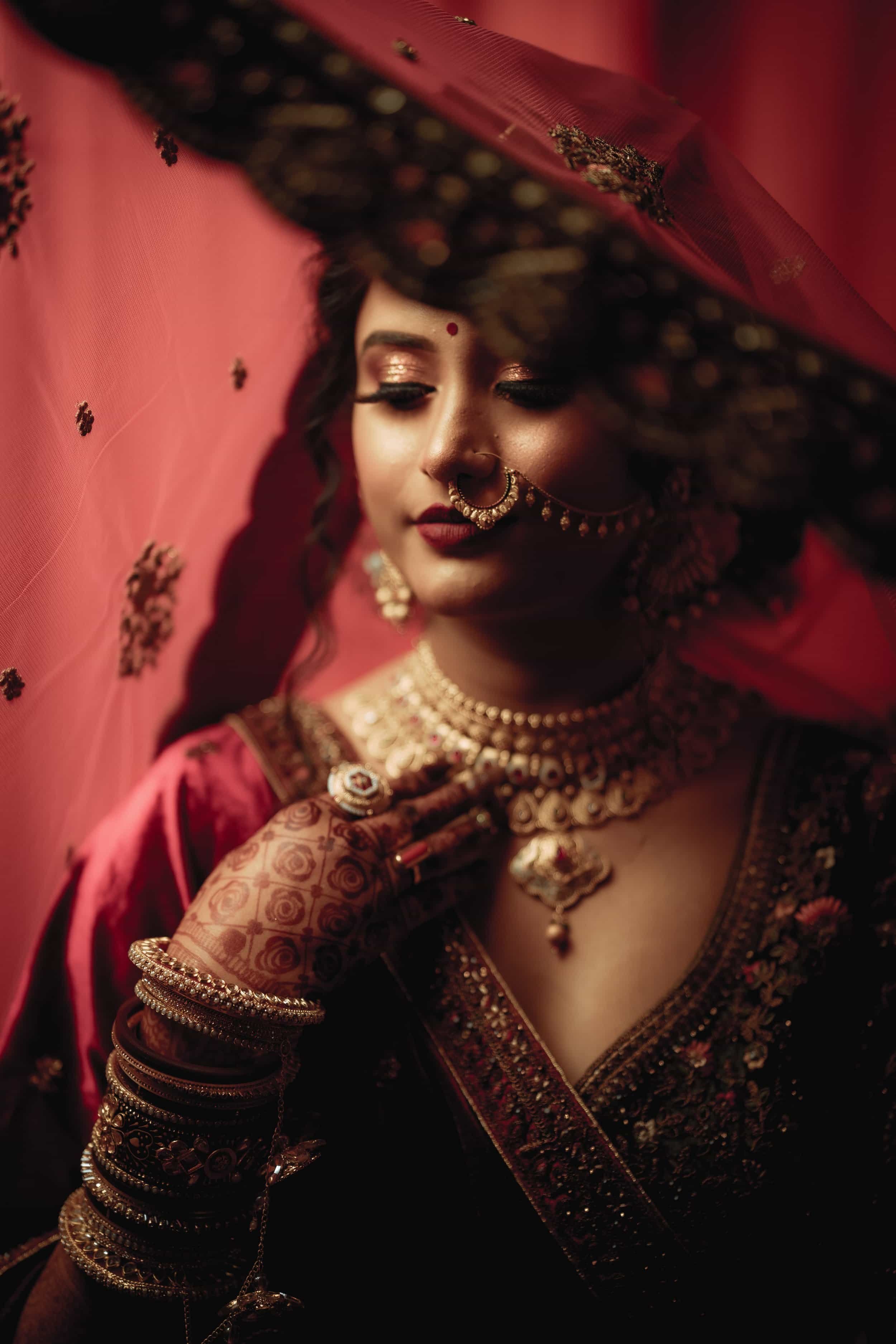 A woman dressed in traditional Indian bridal attire, wearing elaborate jewelry and henna on her hands, with her eyes closed surrounded by red fabric and decorative elements.