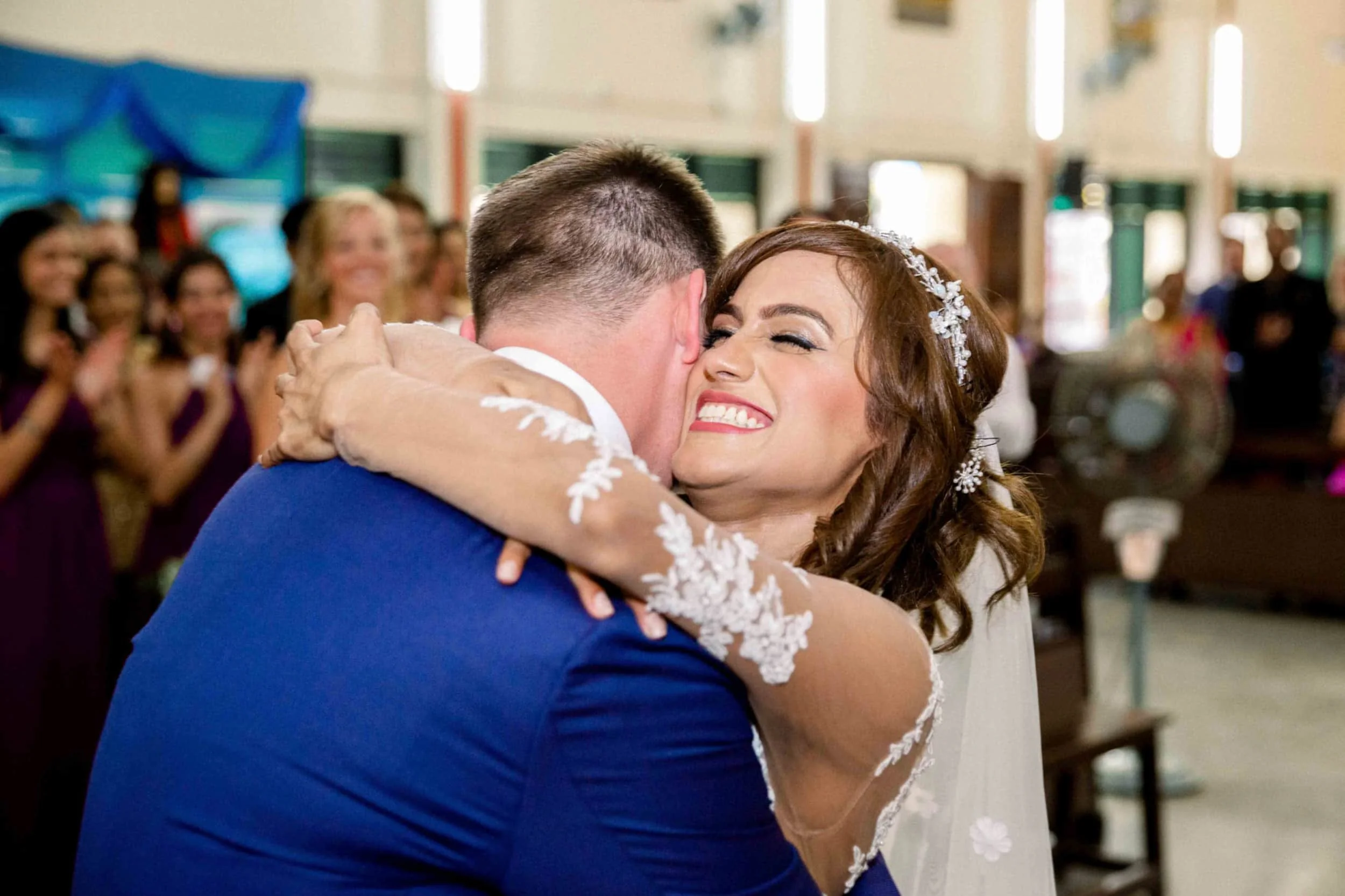 A bride and groom embracing during their wedding ceremony, with guests in the background clapping and smiling.