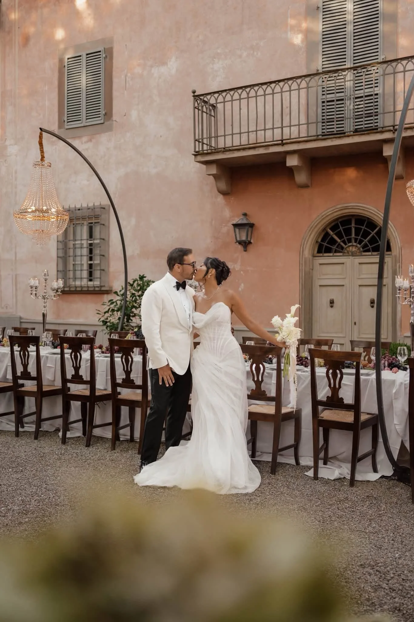 A newlywed couple sharing a kiss at a wedding reception outdoors with elegant chandeliers, a long dining table, and a rustic building with pink walls in the background.