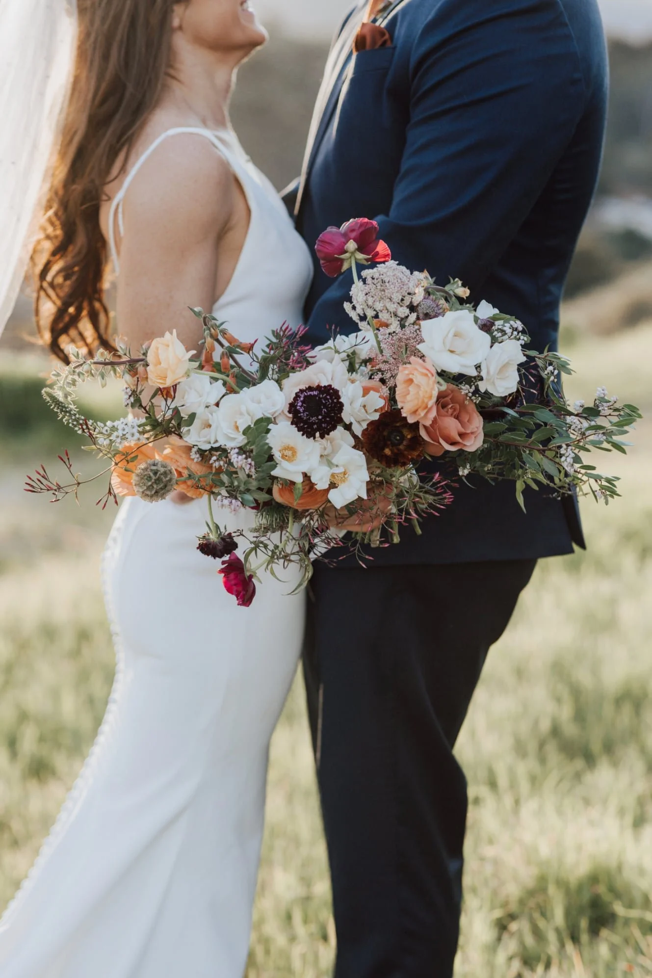 A bride and groom standing close together outdoors, holding a large bouquet of mixed flowers including roses and poppies.
