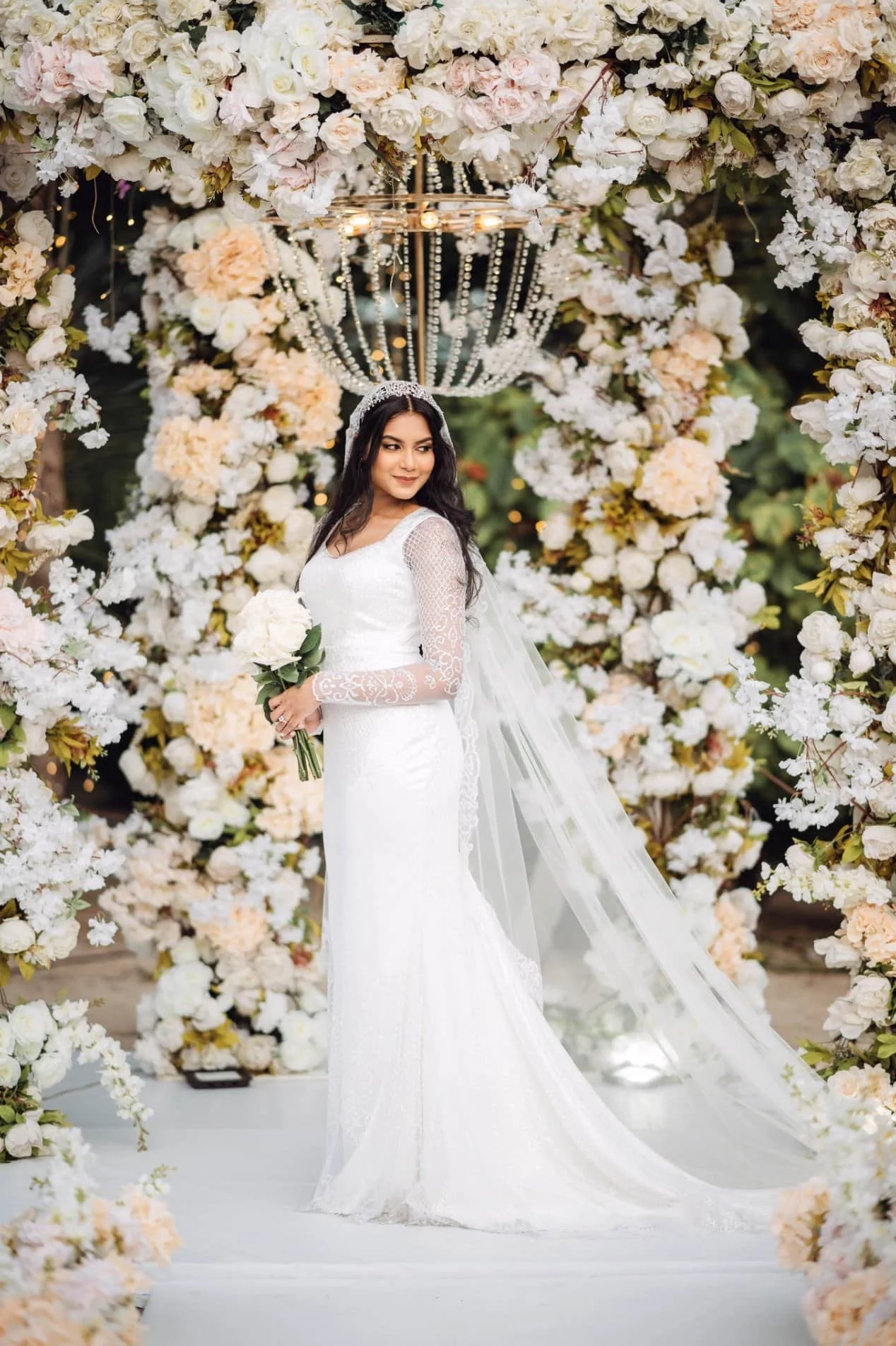 A bride in a white wedding dress holding a bouquet of white flowers, standing in a floral archway decorated with white and light pink flowers and a chandelier overhead.