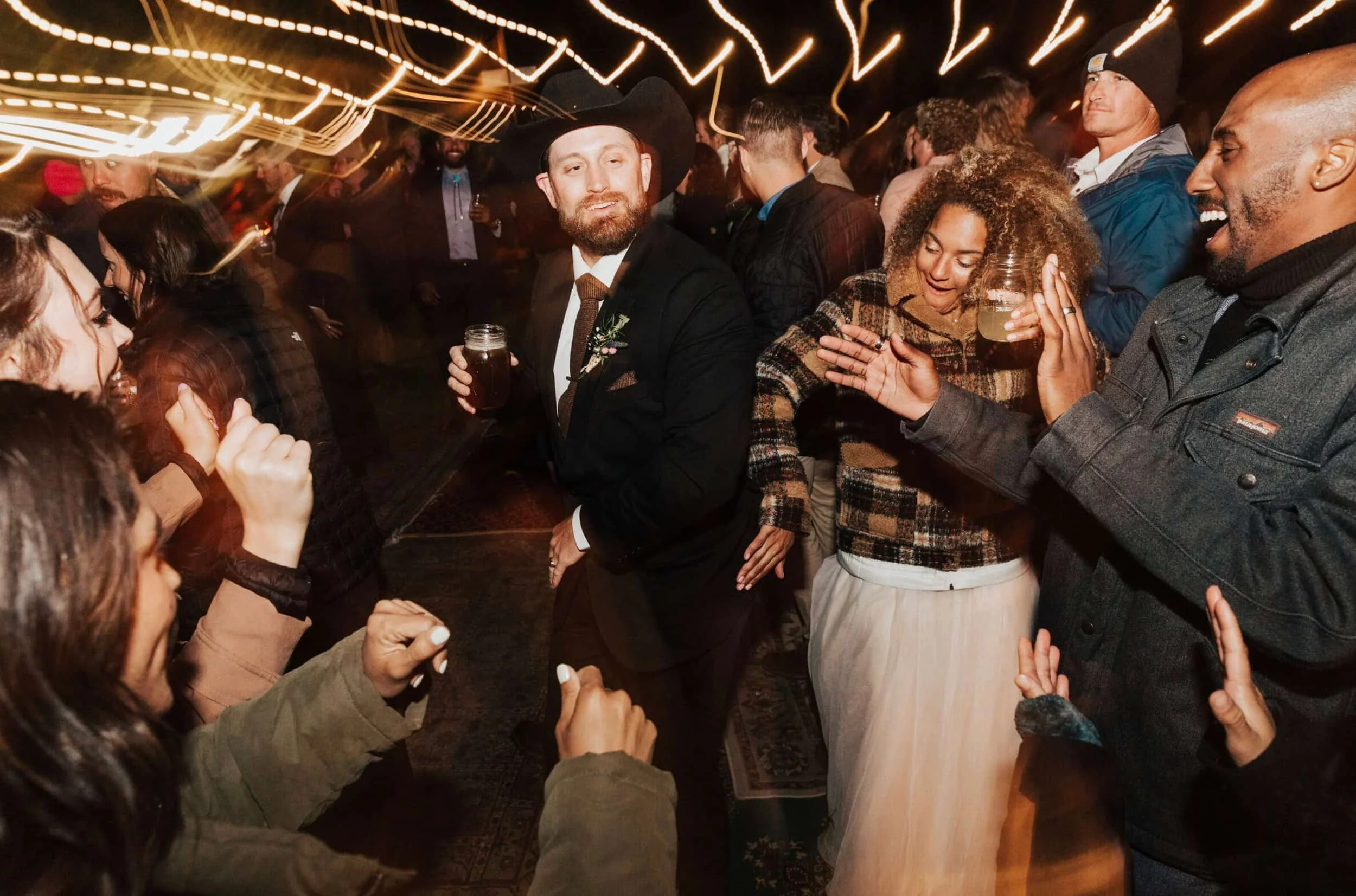 People socializing at a party under string lights, with a man in a cowboy hat and suit holding a drink in the center.