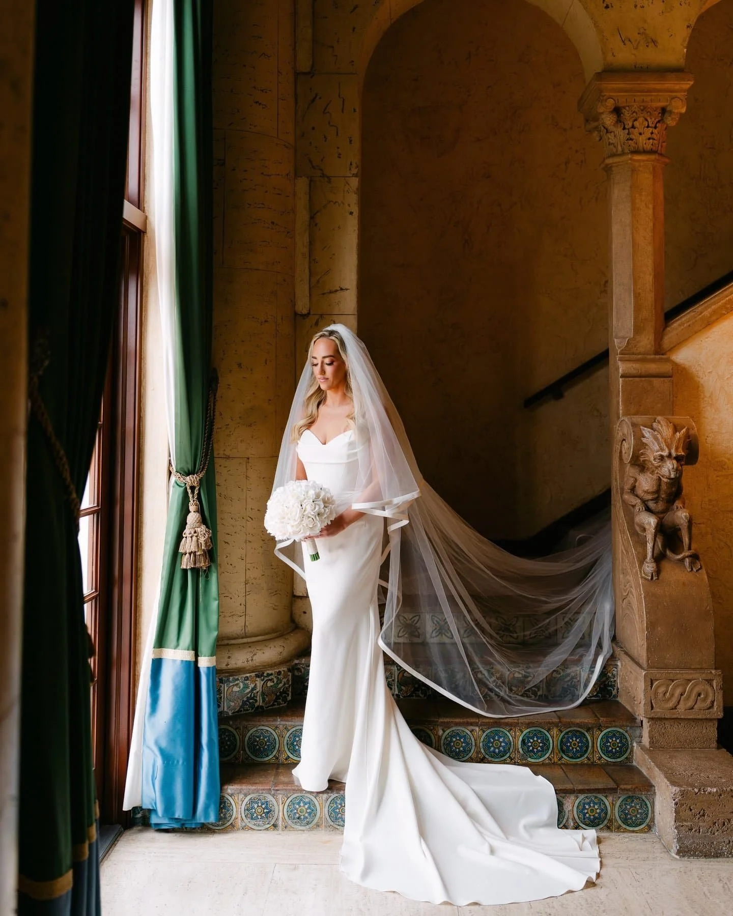 A bride in a white wedding dress holding a bouquet of white flowers, standing by a window with blue and green curtains and looking down, inside a historic building with ornate stone columns and carved sculptures.