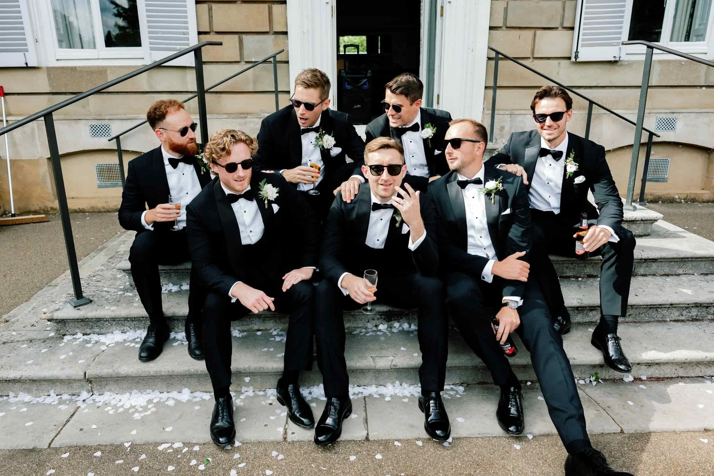 Group of men in tuxedos and sunglasses sitting and standing on steps outside a building, celebrating with drinks at a wedding or formal event.