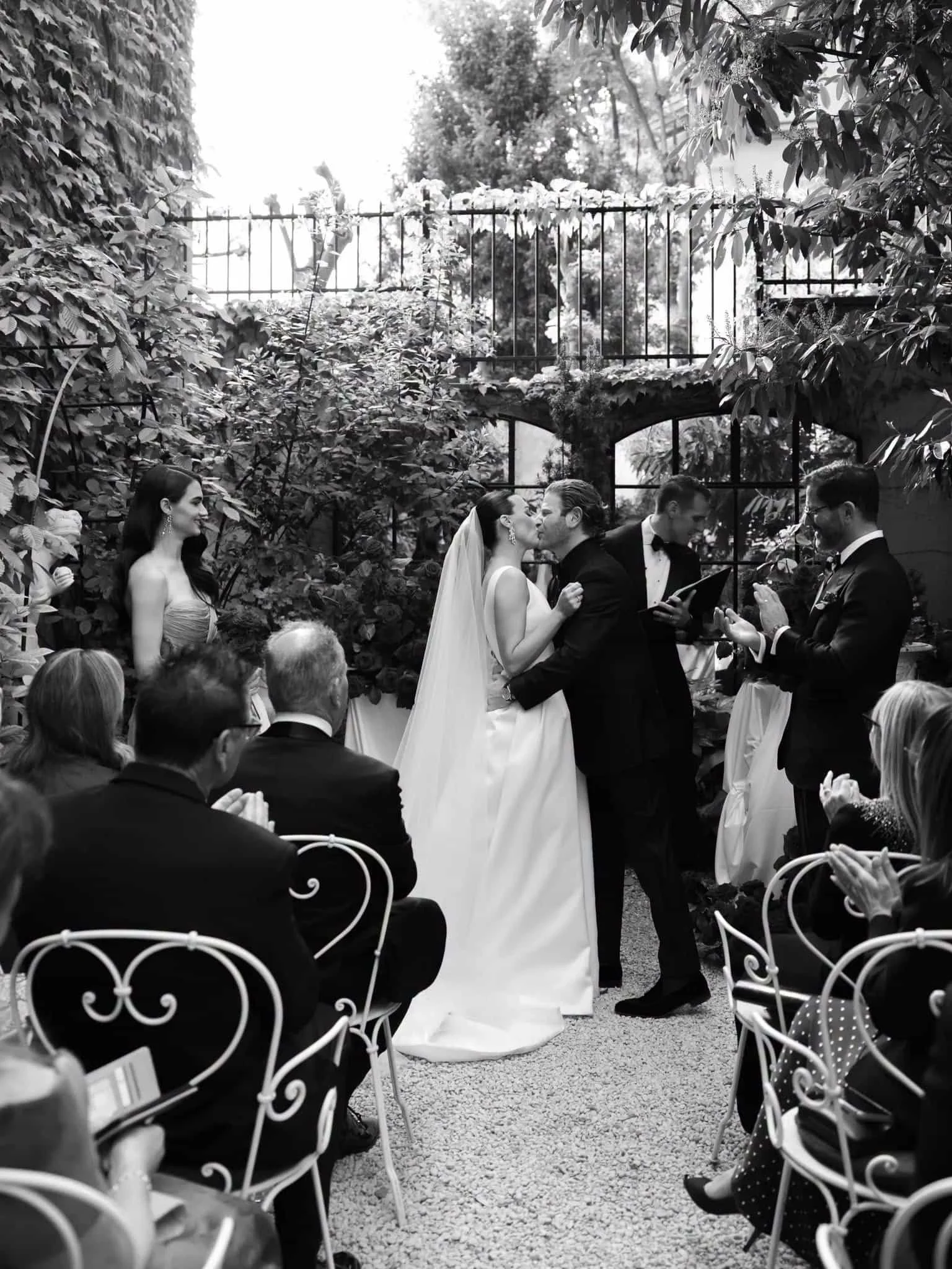 A wedding ceremony taking place in a garden with lush plants and a large window. A bride and groom are kissing, while their wedding officiant reads from a book. Several guests are seated, watching the ceremony.