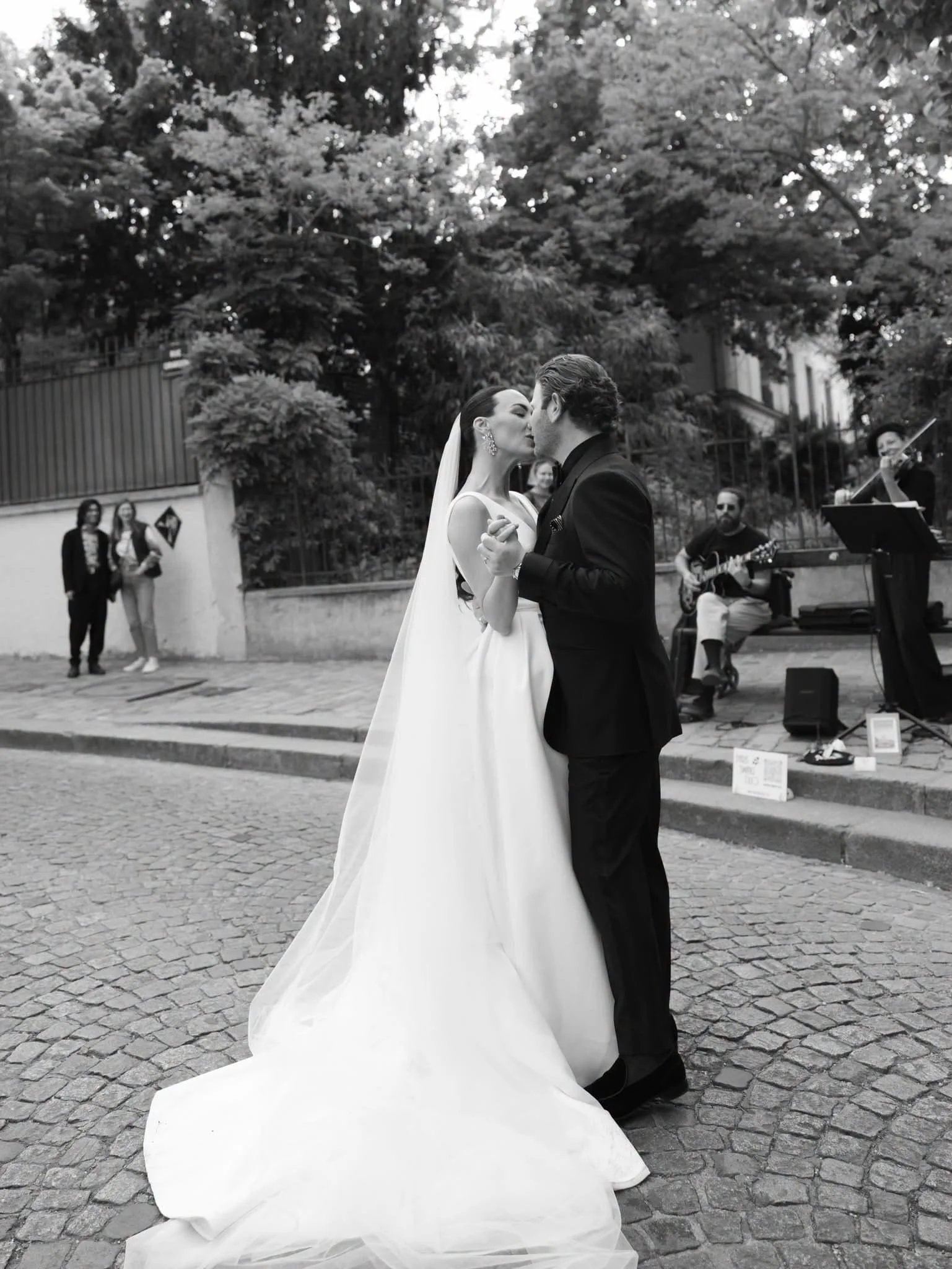 A bride and groom are dancing outdoors during their wedding, with musicians playing in the background and a few guests watching.