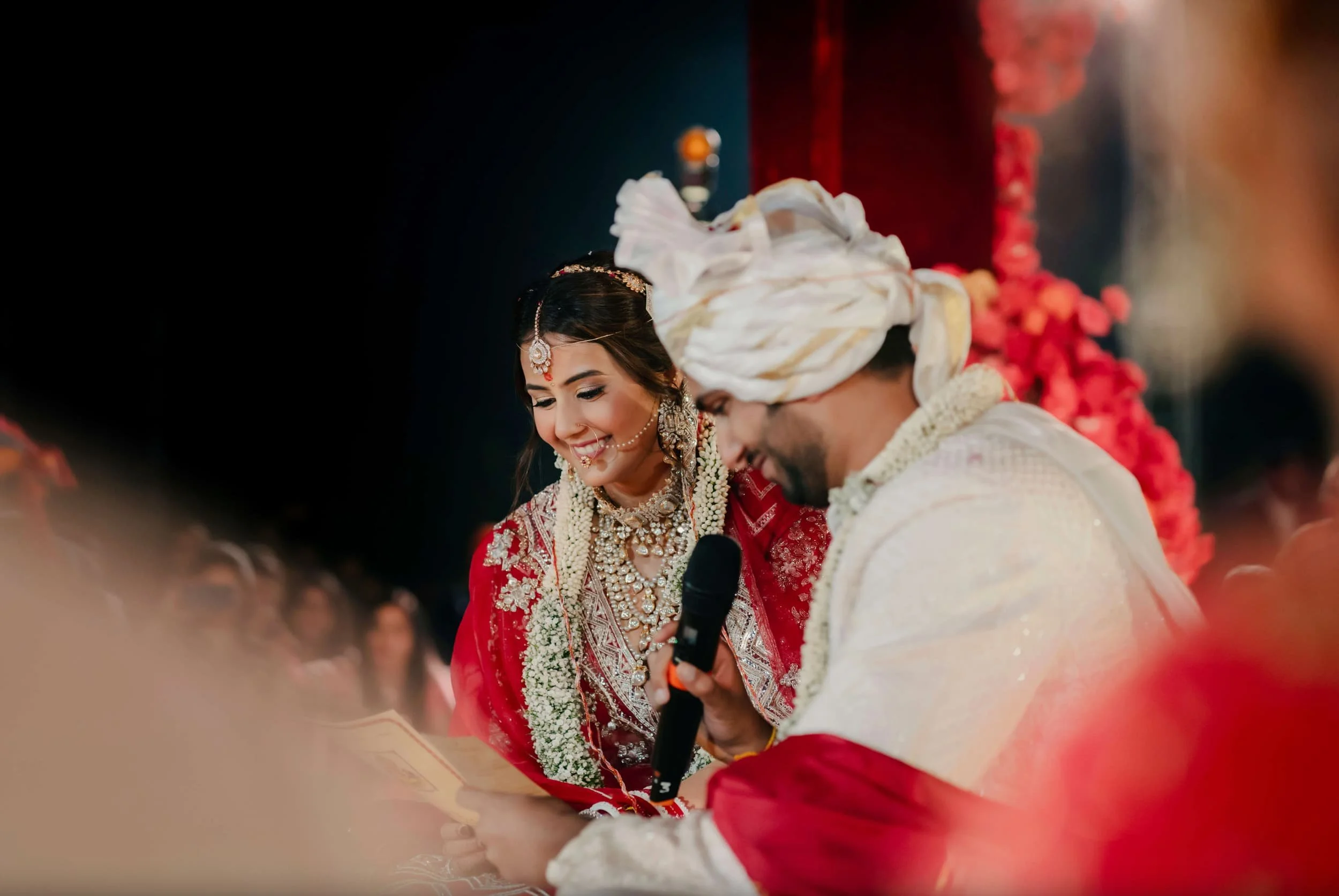 Indian bride and groom during wedding ceremony, dressed in traditional attire, with flower decorations in the background.