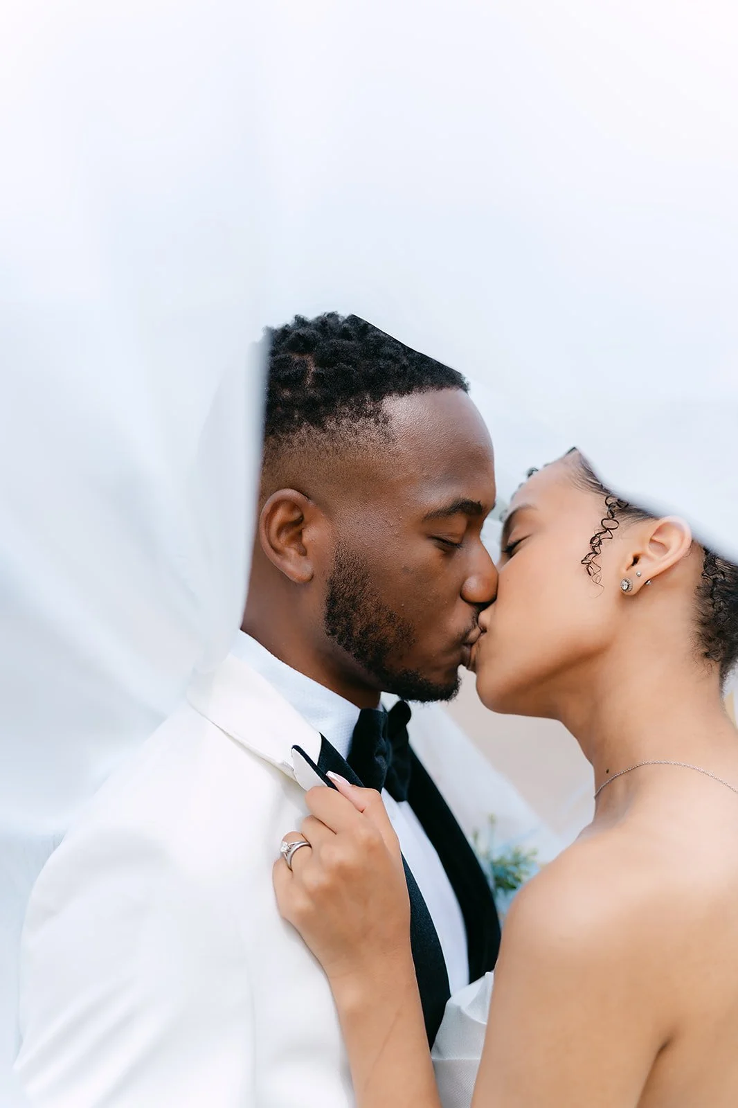 A couple sharing a kiss, dressed in wedding attire against a white background, with the bride wearing earrings and a necklace.