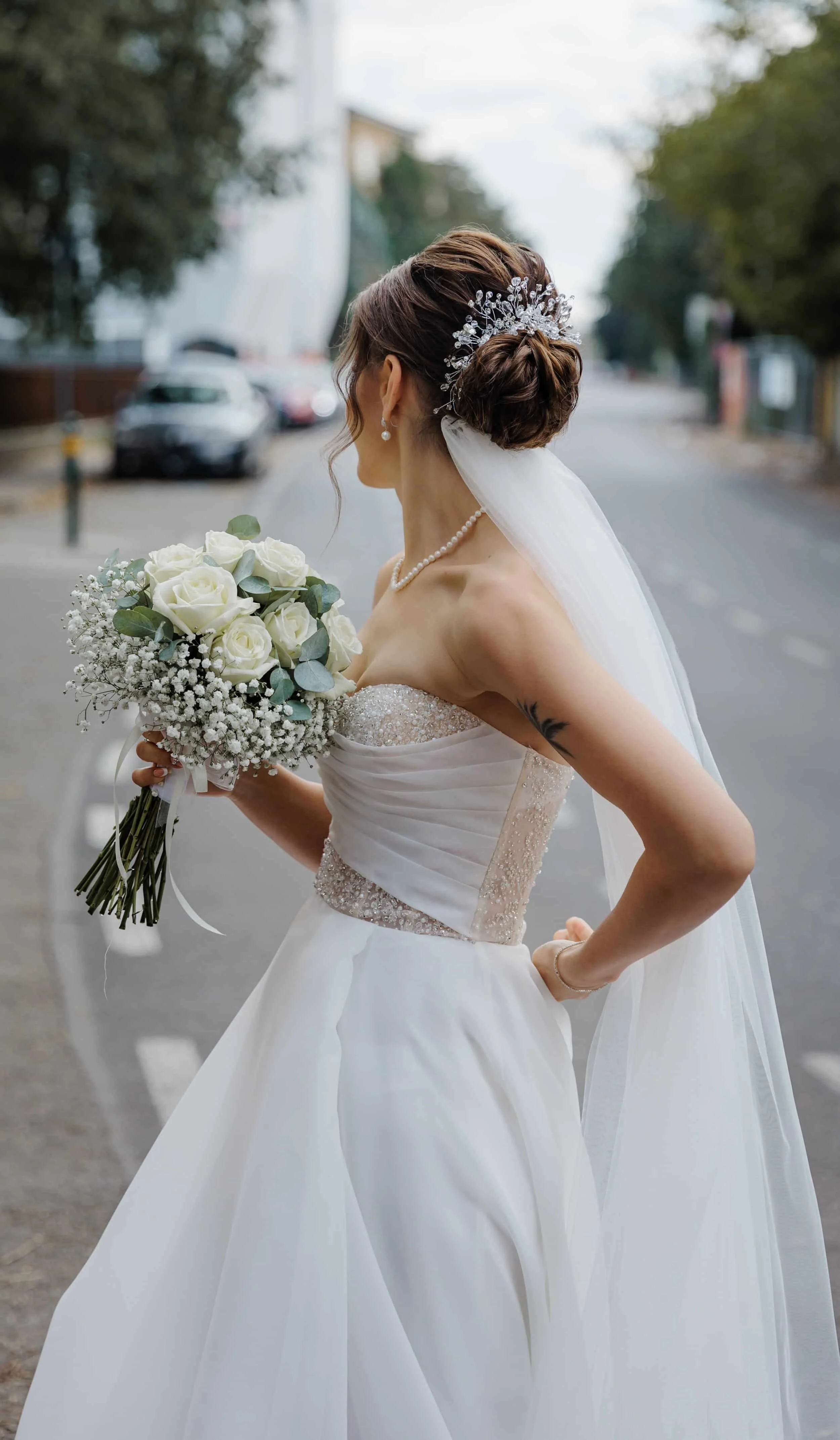 A bride with a wedding dress, pearl jewelry, and a tiara, holding a bouquet of white roses and baby's breath, standing outdoors with trees and parked cars in the background.