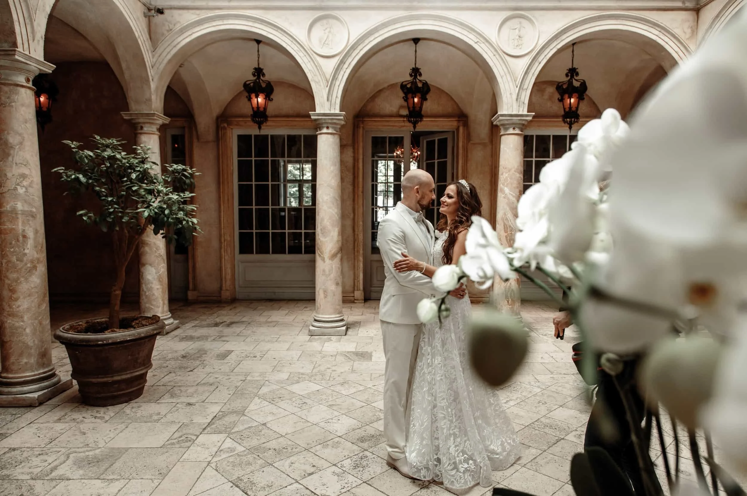 A bride and groom share a moment in an elegant indoor wedding venue with arched ceilings, large windows, and ornate lighting. The bride is wearing a lace wedding dress with a tiara, and the groom is dressed in a white suit. There are potted plants an