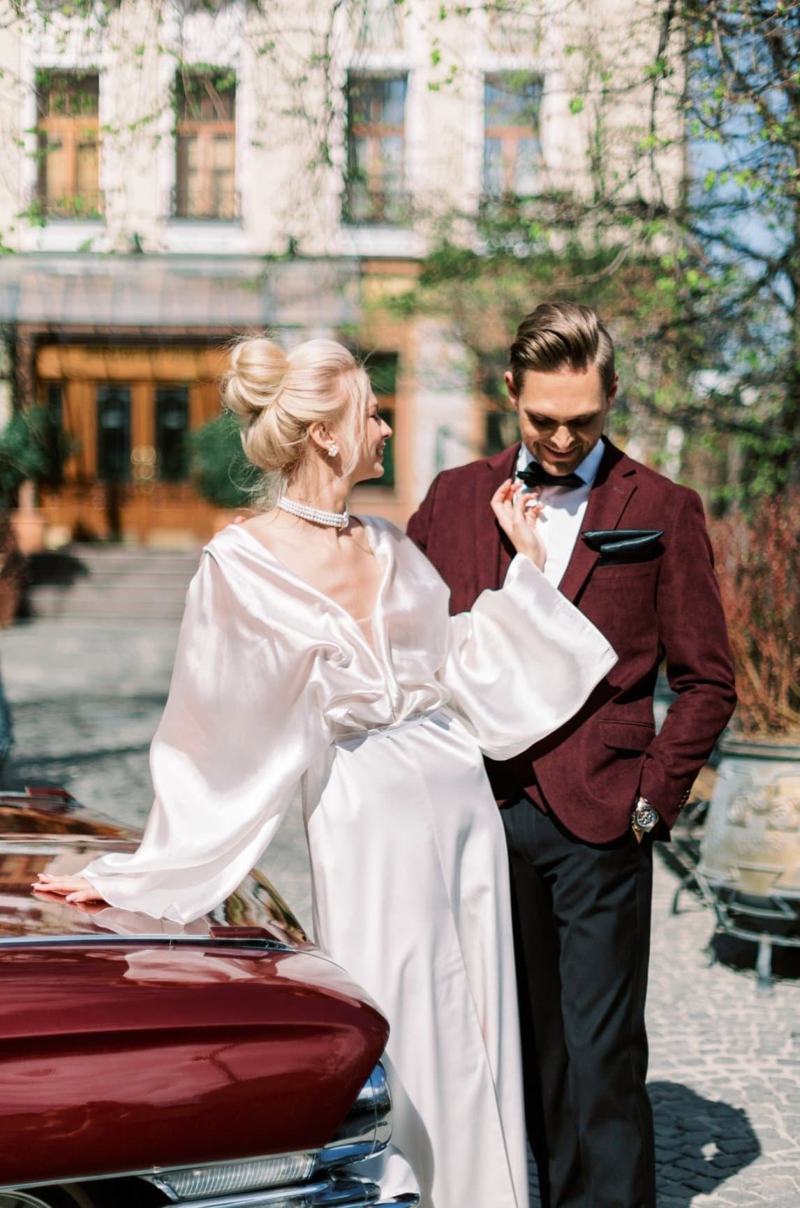 A woman in a white satin dress with long, flowing sleeves and pearl necklace, smiling and laughing, interacting with a man in a burgundy suit jacket, white shirt, and black bow tie, outdoors on a sunny day with a tree and building in the background.